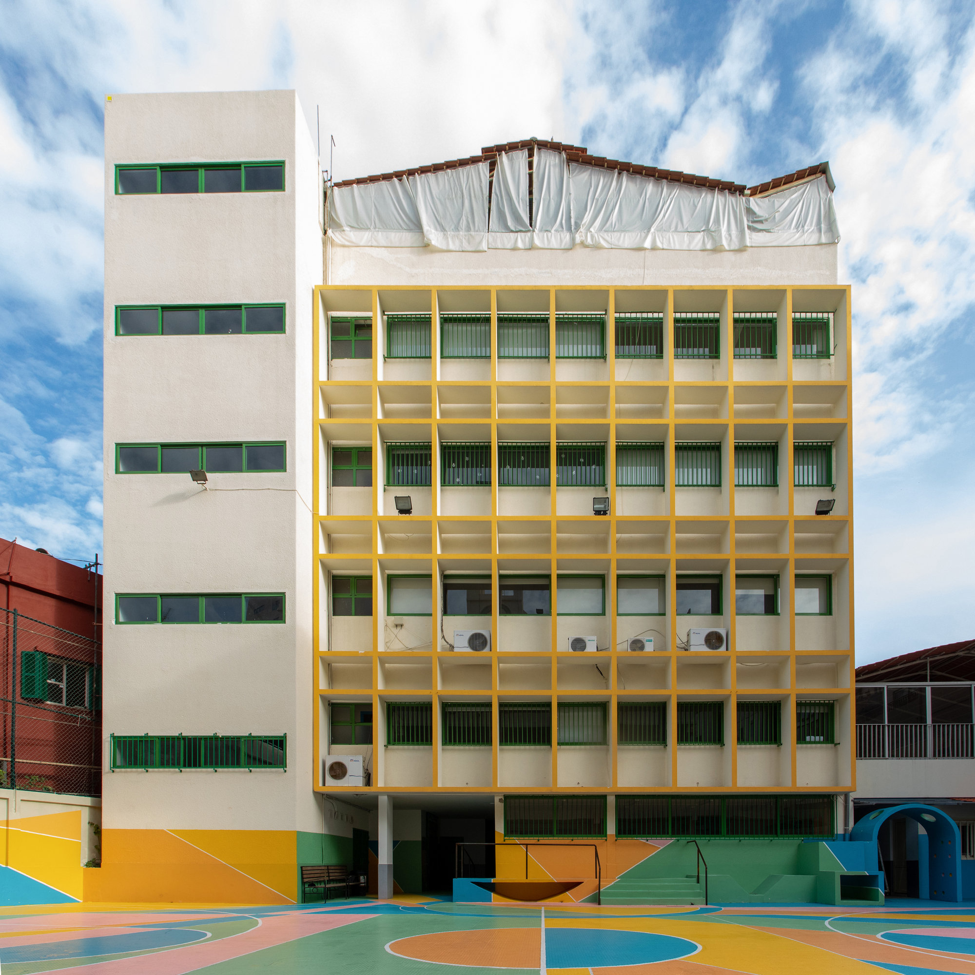 Yellow-framed concrete facade with horizontal windows beside white tower under partly cloudy sky