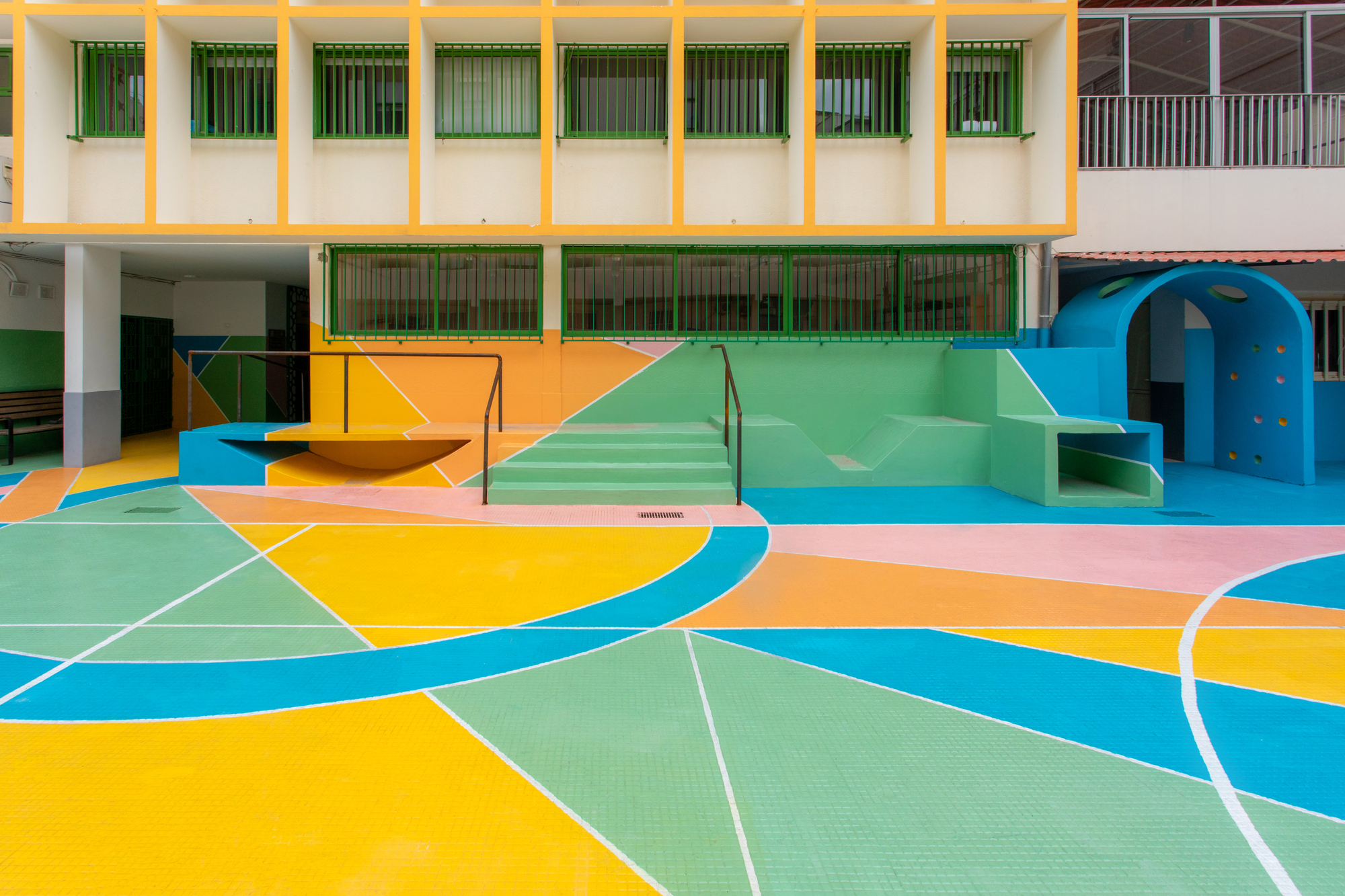 Painted concrete stairs and play structure on pastel geometric courtyard surface below yellow-framed classroom wing