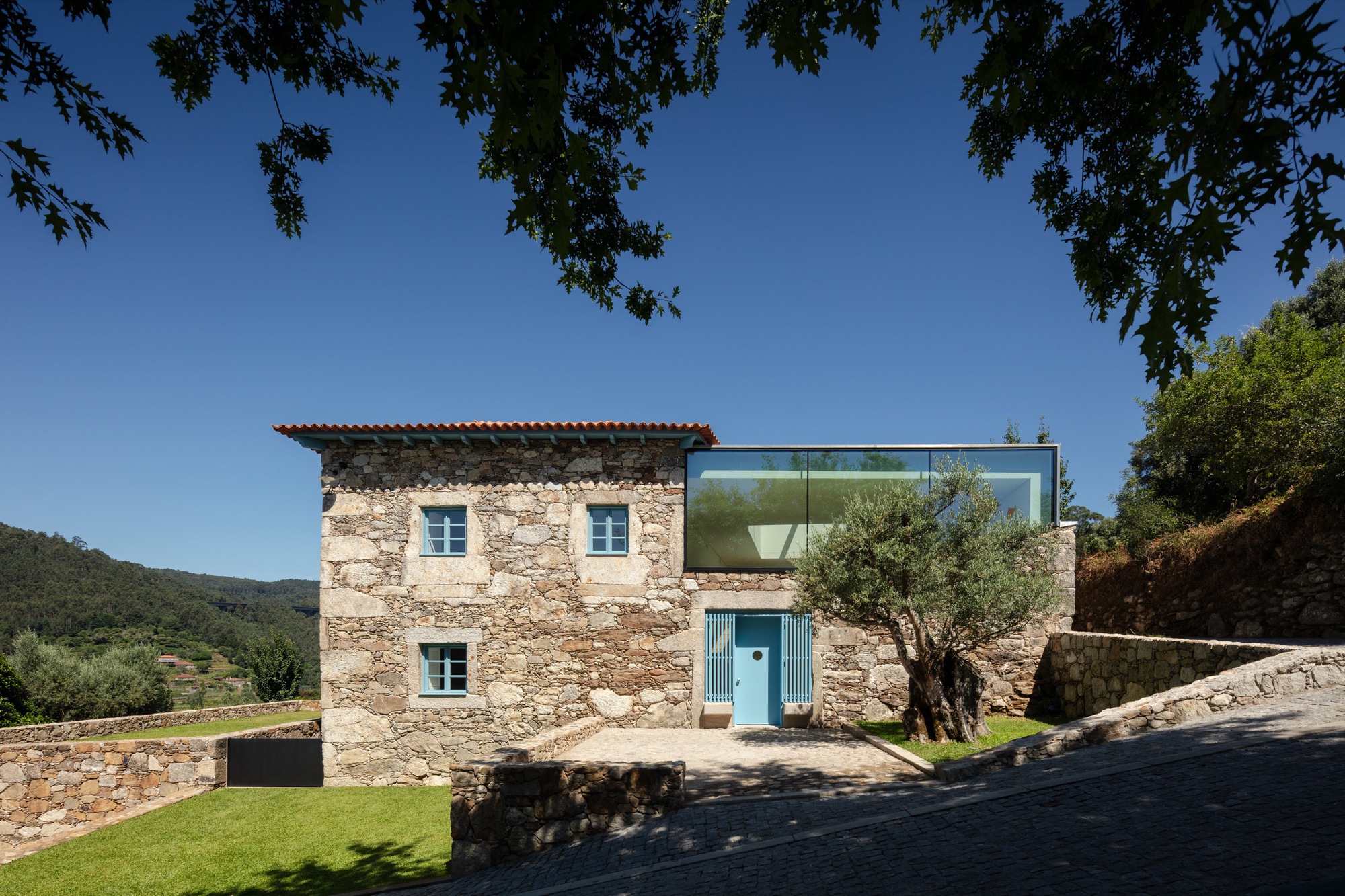 Stone farmhouse with blue shuttered windows and glass addition set into terraced lawn under clear sky