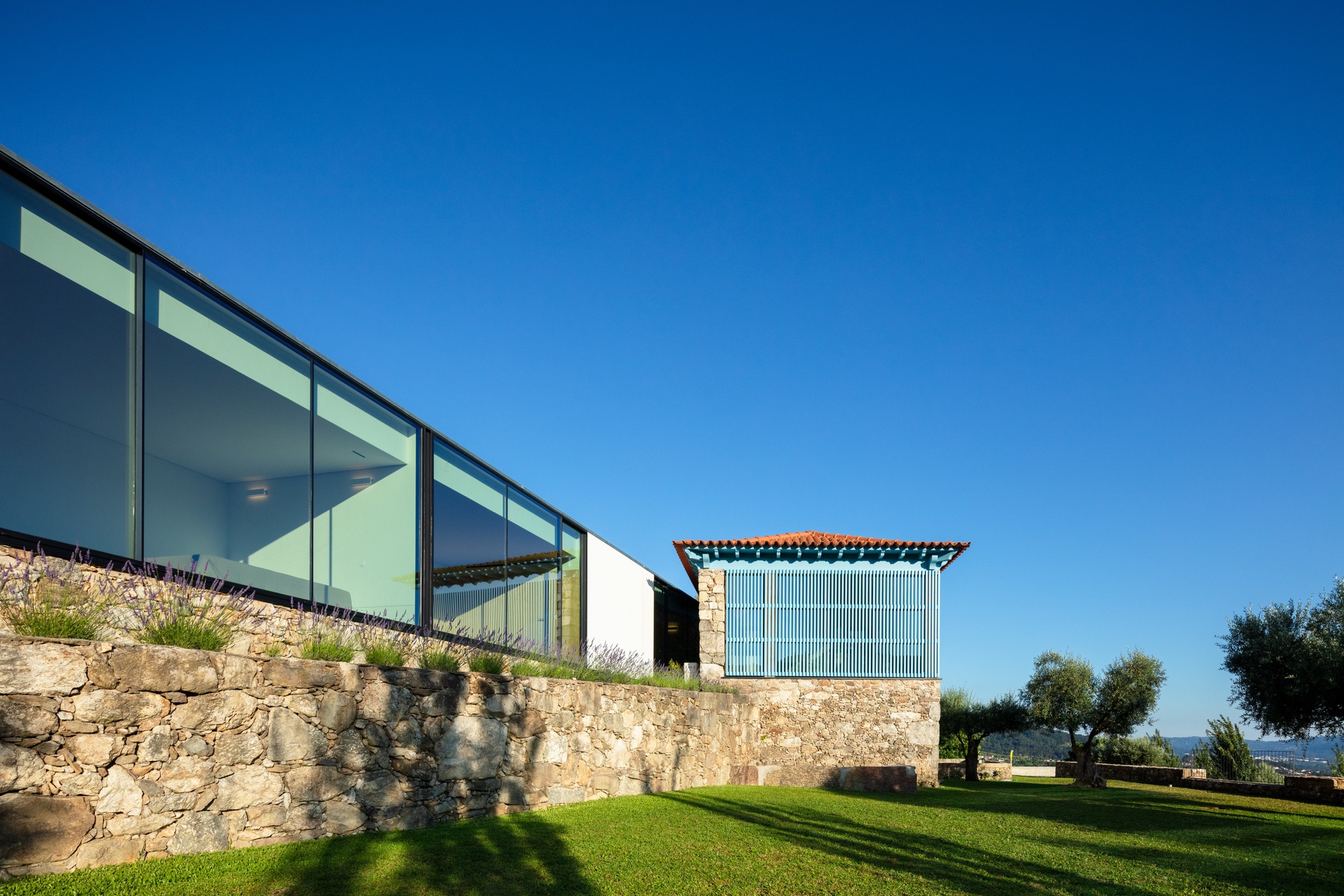 Glass pavilion extension above stone retaining wall with lawn stretching toward distant olive trees