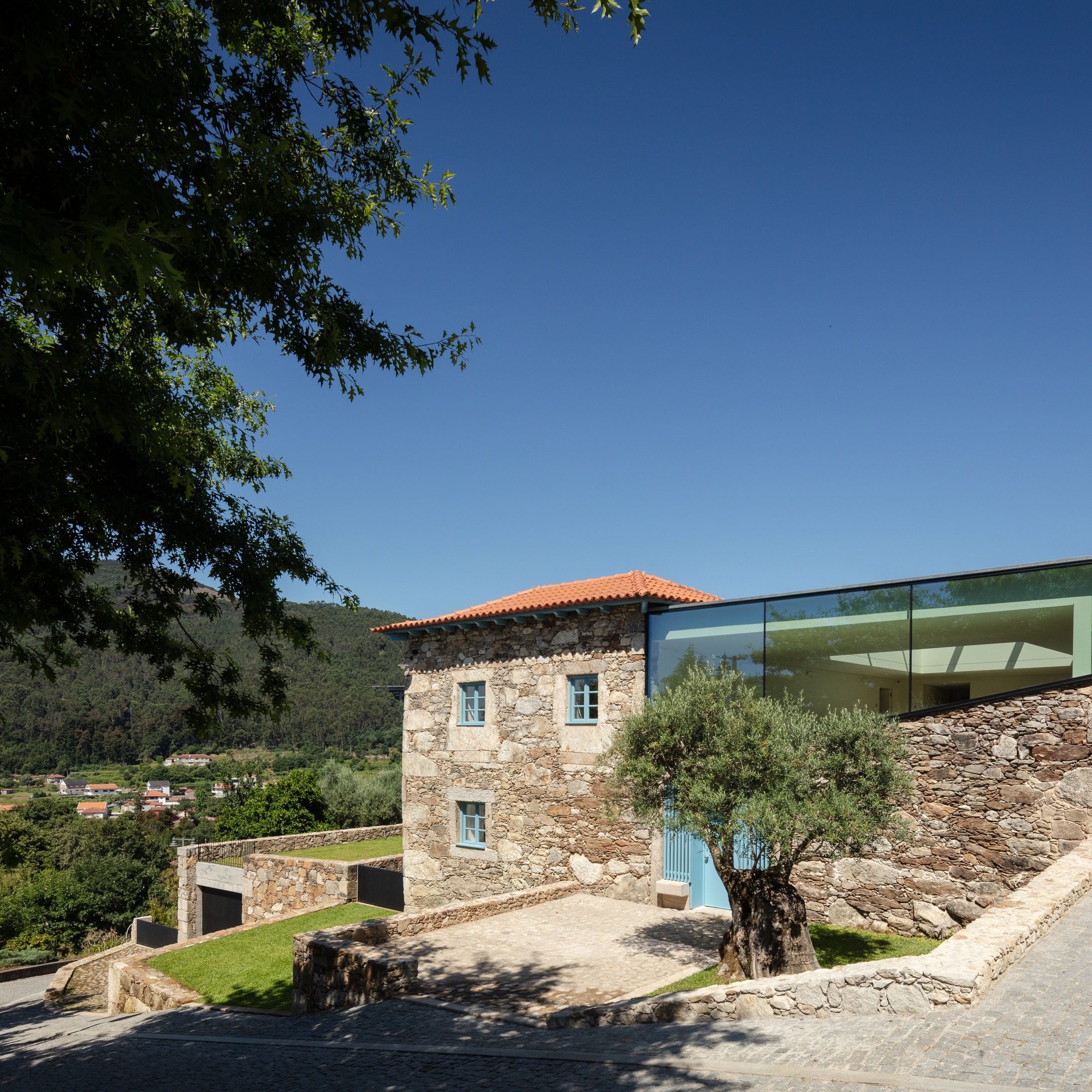 Stone tower with tiled roof beside glass volume framed by olive tree and overhanging branches