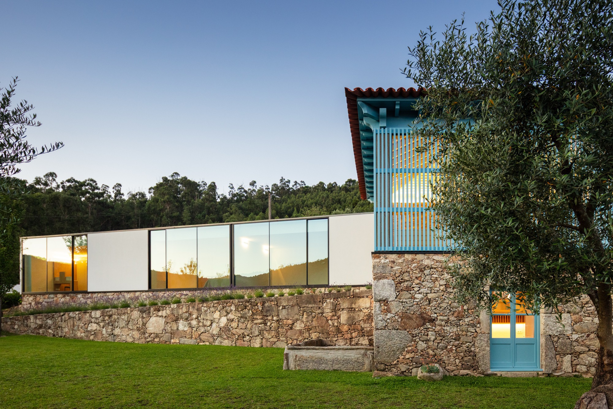 Garden facade with illuminated interiors behind corrugated blue tower and stone base at dusk
