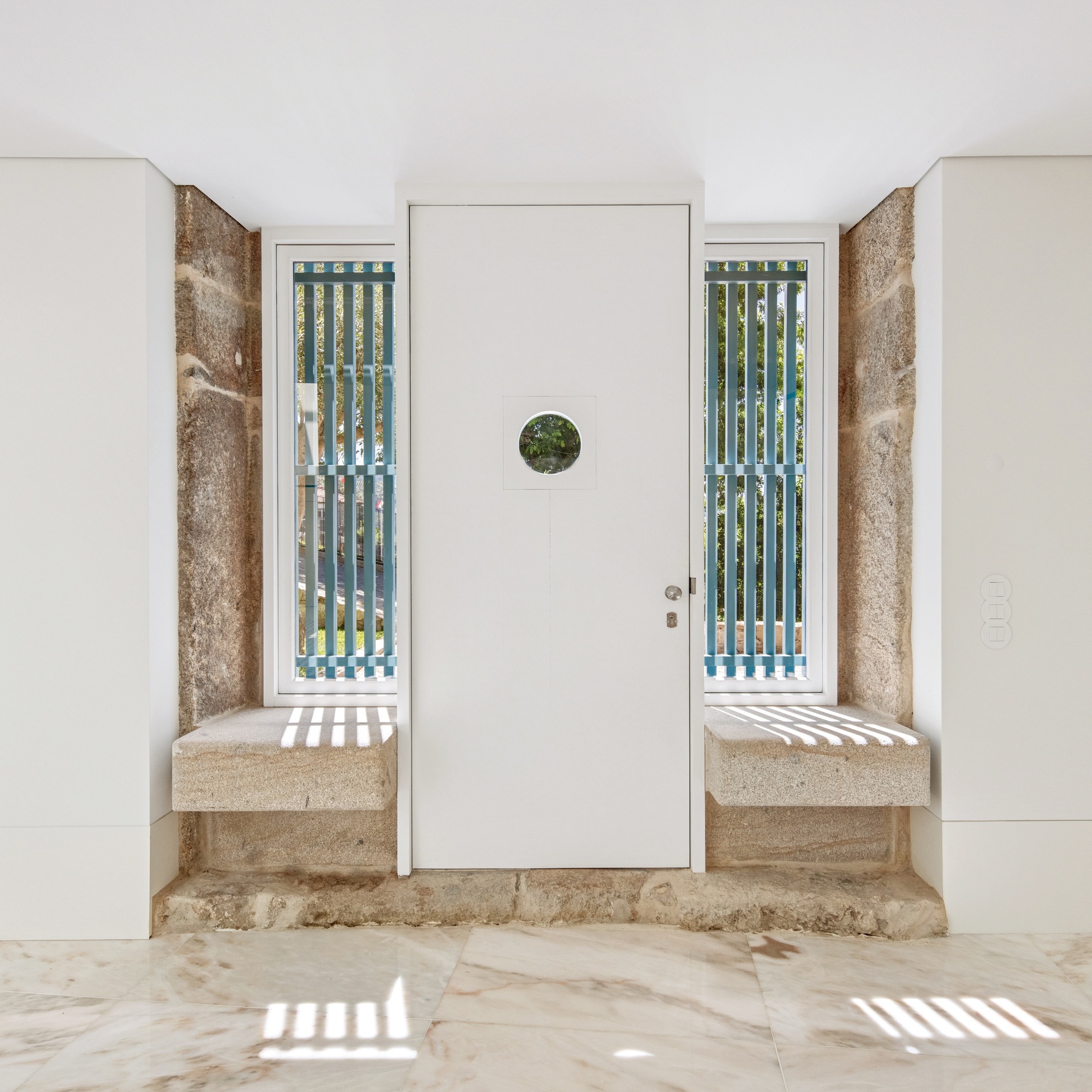 White door with circular window framed by weathered stone reveals and vertical slatted sidelights casting shadow stripes