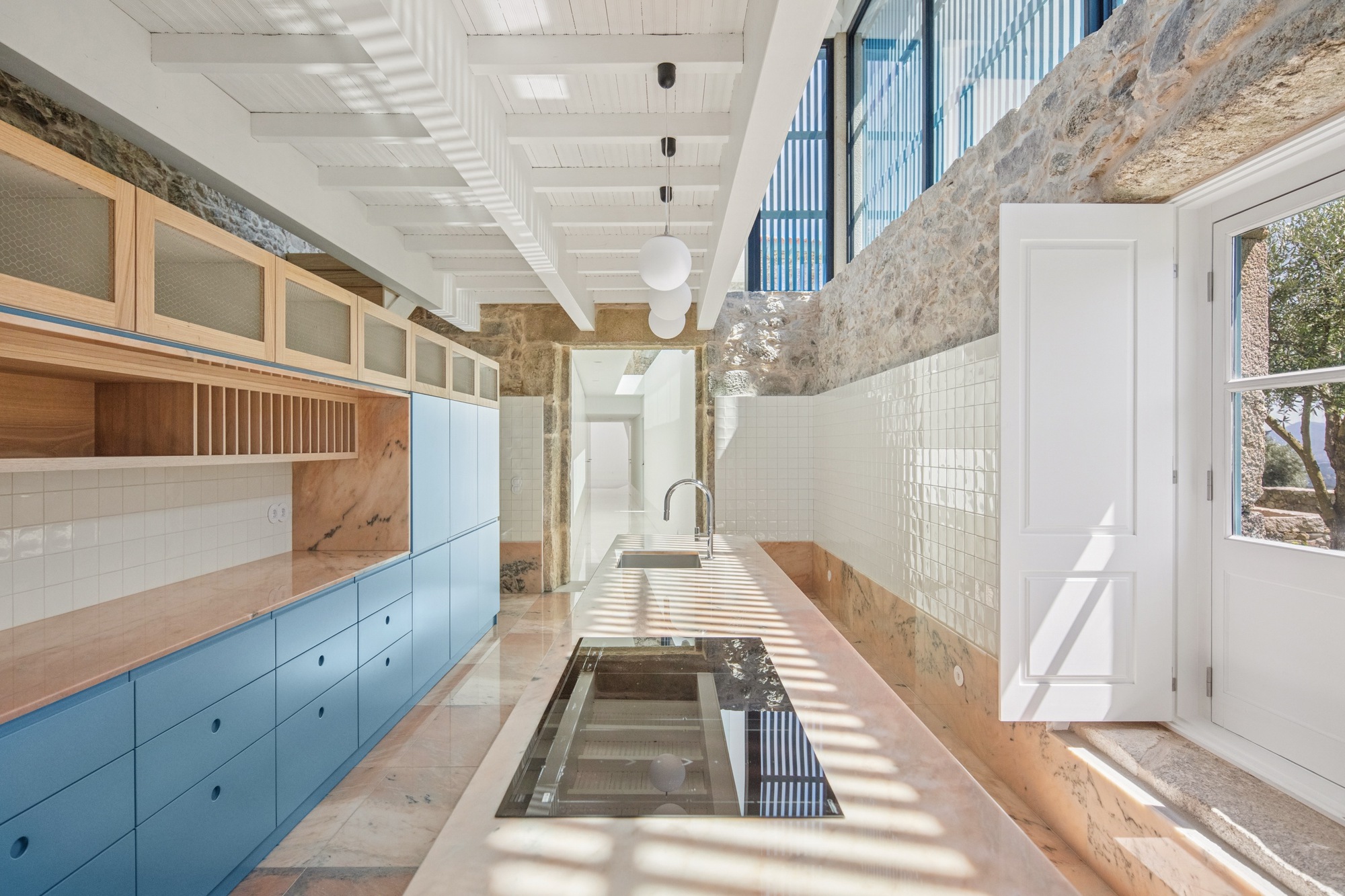 Kitchen with blue timber cabinetry, exposed stone walls, white painted beams and vertical louvres filtering daylight