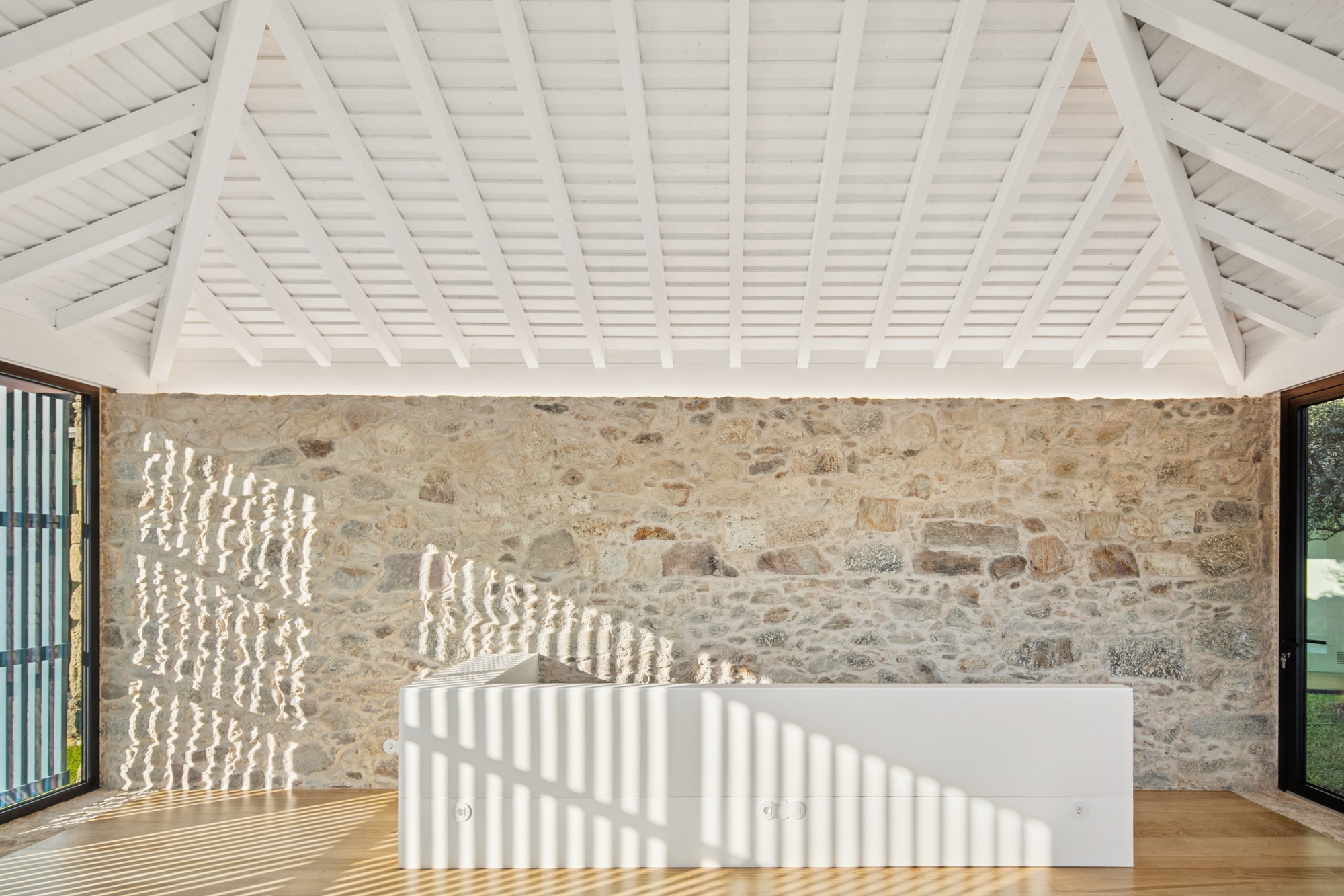 Interior with exposed stone wall and white painted timber ceiling casting striped shadows across wooden floor