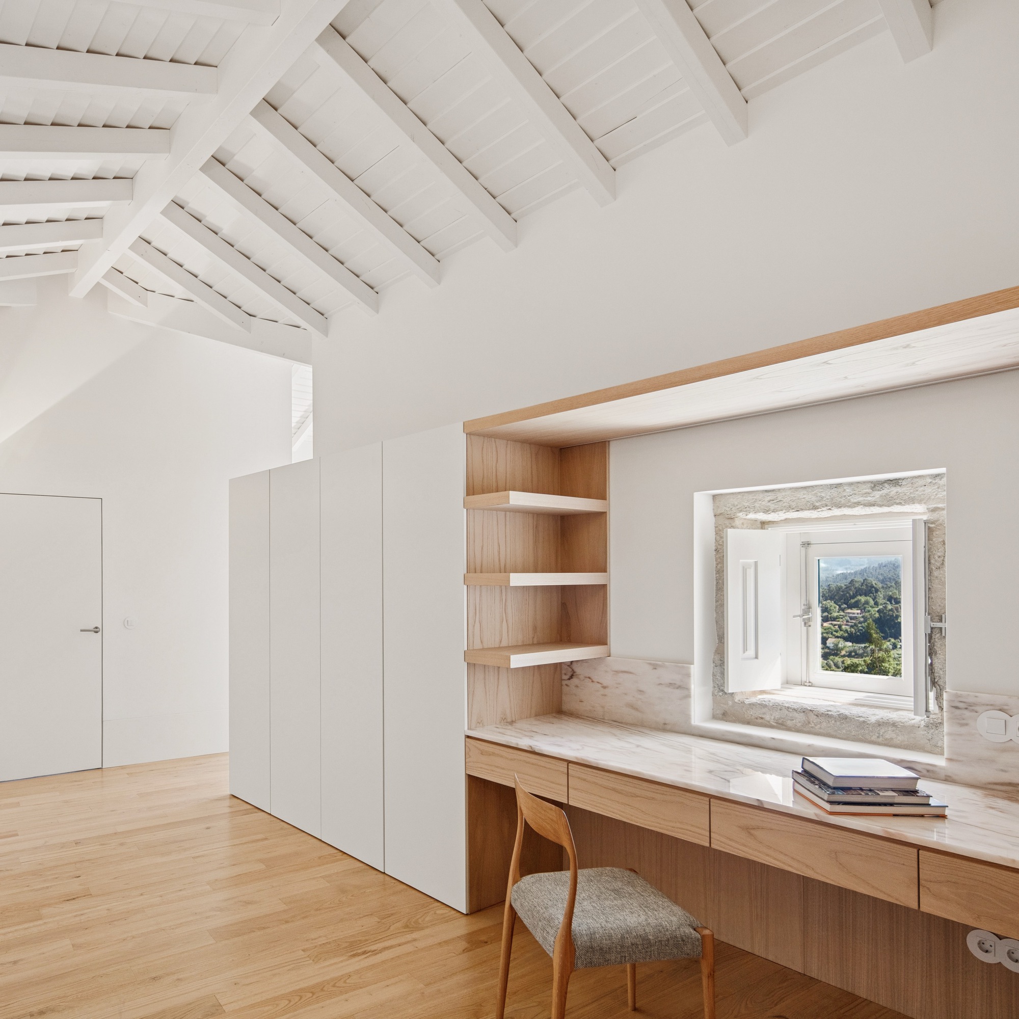 Built-in timber desk with shelving beneath exposed white rafters framing a deep stone window opening
