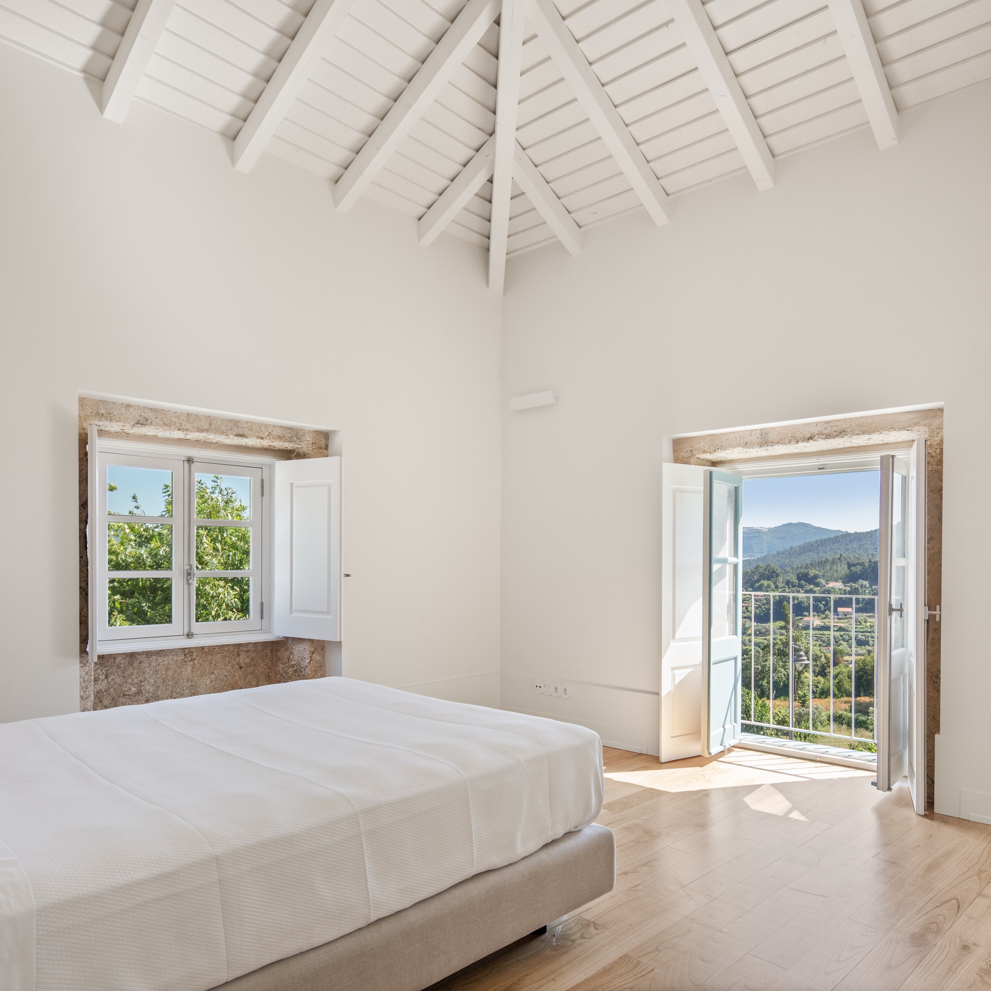Bedroom with pitched white ceiling and two stone-framed openings overlooking hillside landscape in daylight