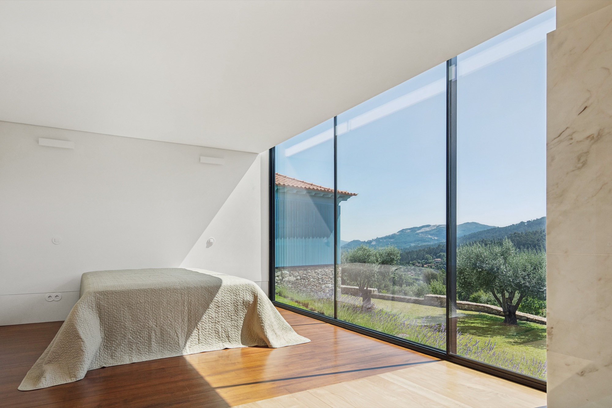 Bedroom with floor-to-ceiling glazing framing view of olive grove and distant hills under clear sky