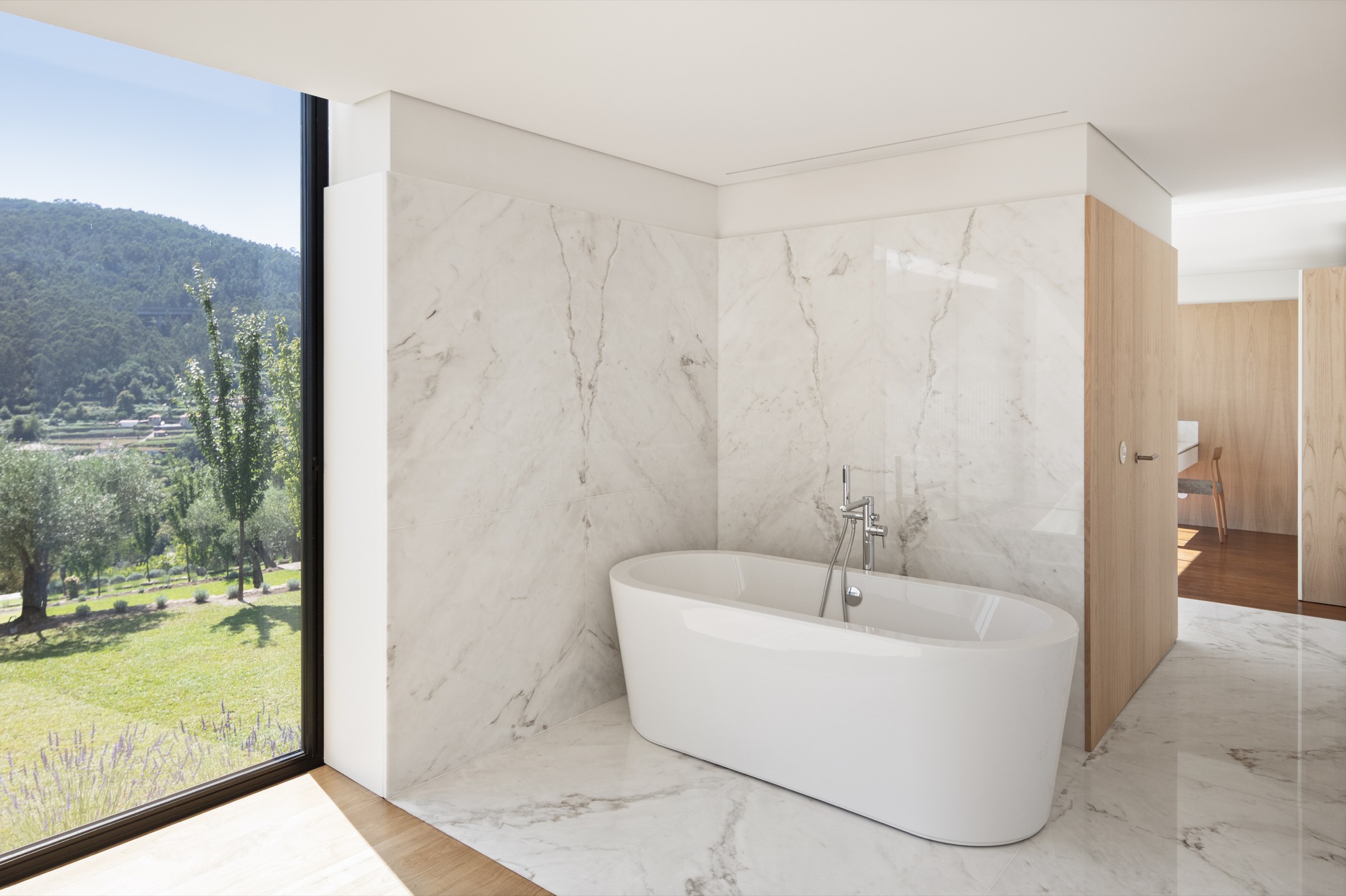 Freestanding bathtub enclosed by white marble walls with floor-to-ceiling window overlooking olive trees