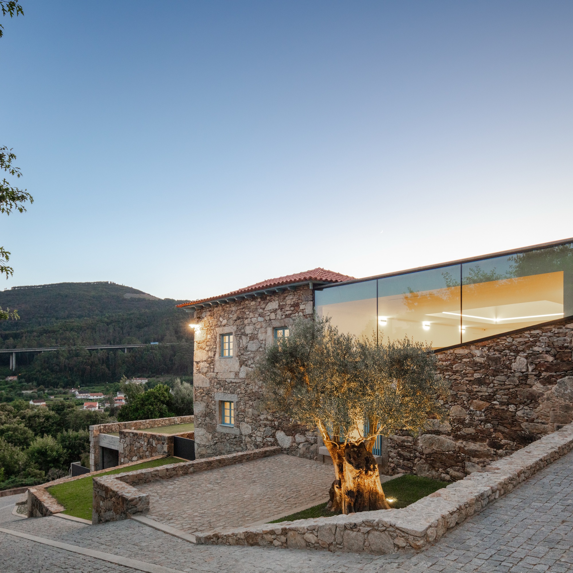 Stone tower and glass volume overlooking a terraced courtyard with olive tree at dusk