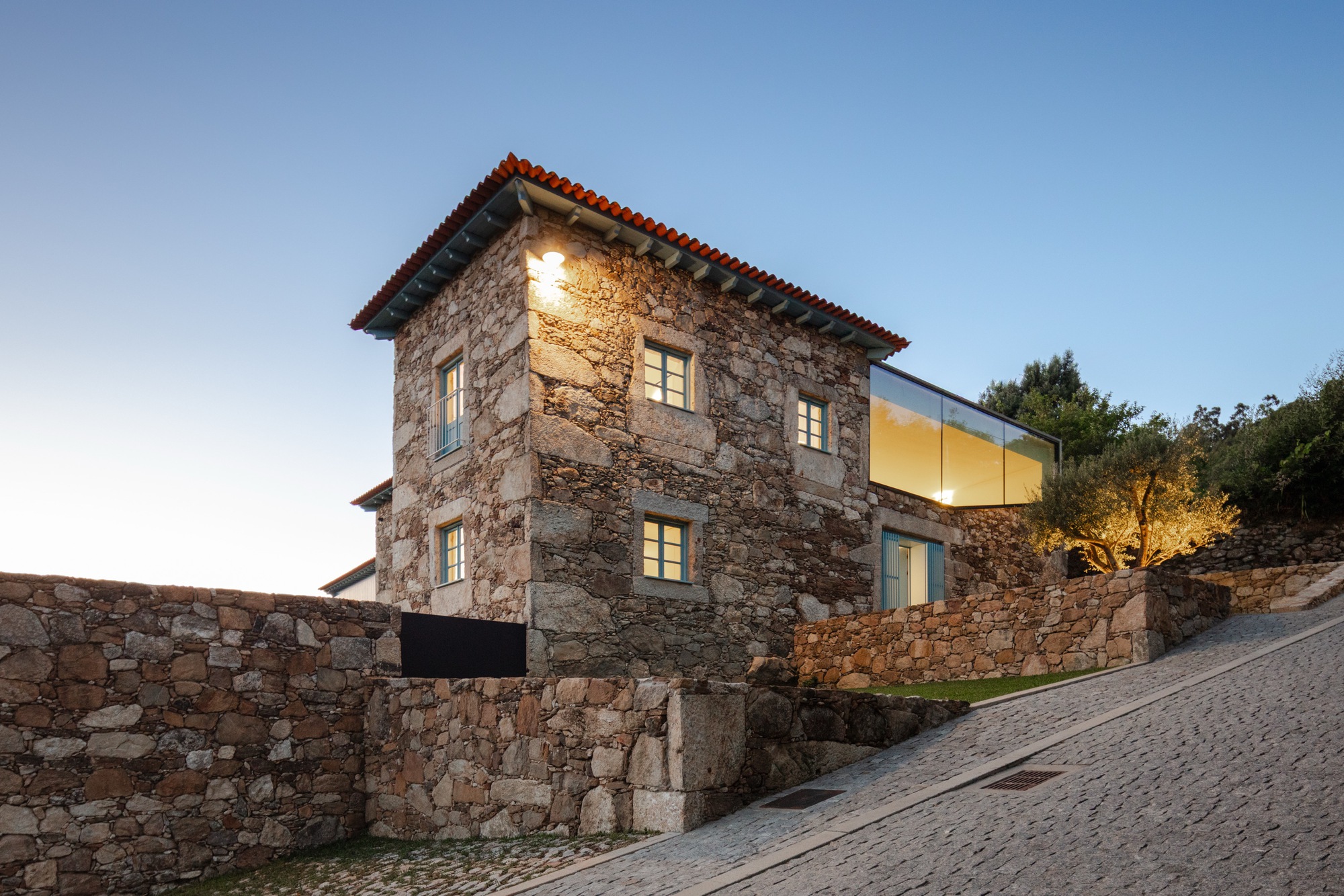 Stone tower with terracotta roof adjacent to glass extension above retaining wall at twilight