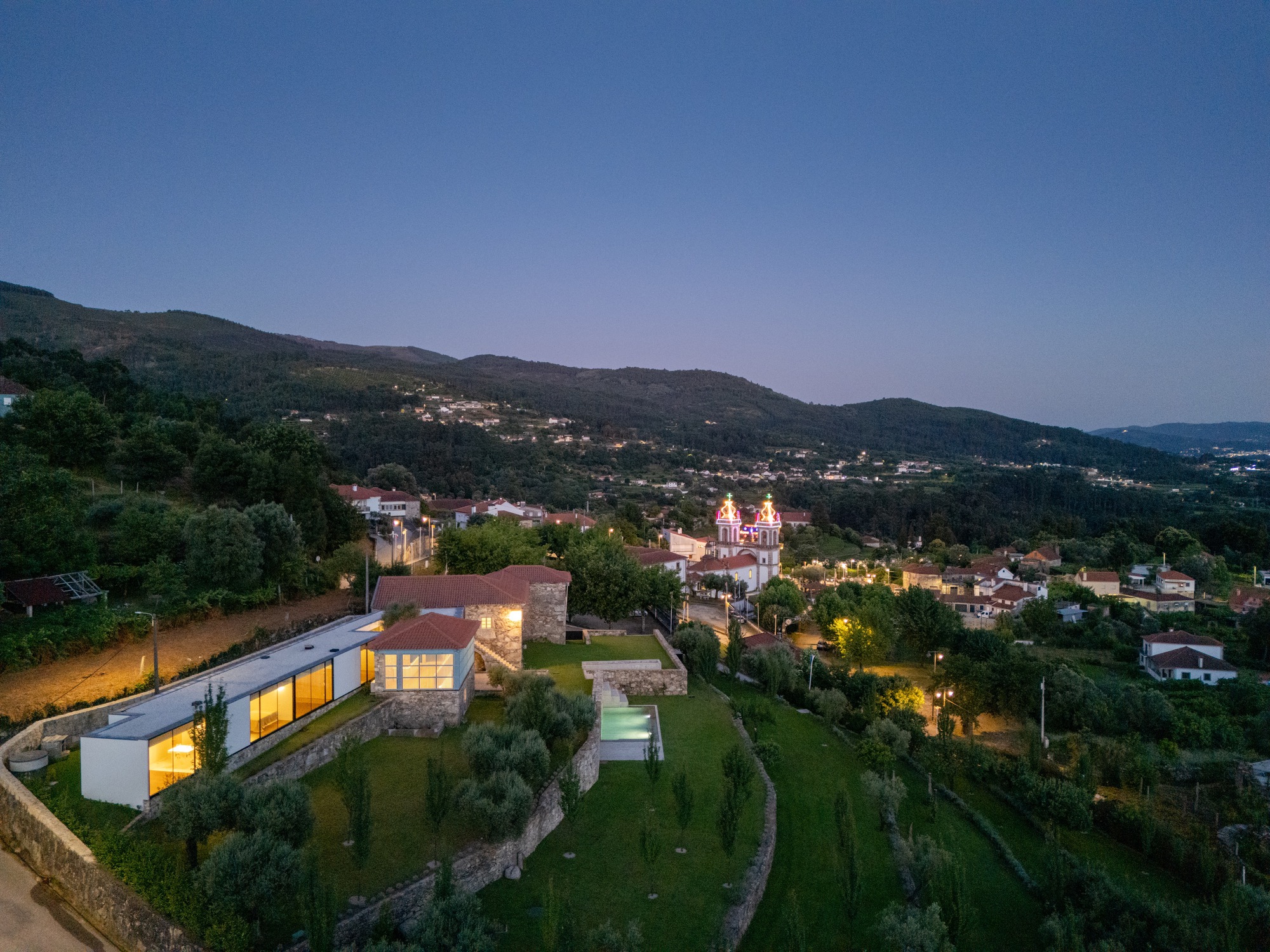 Aerial view of stone tower and glass volumes on hillside terraces with pool at dusk