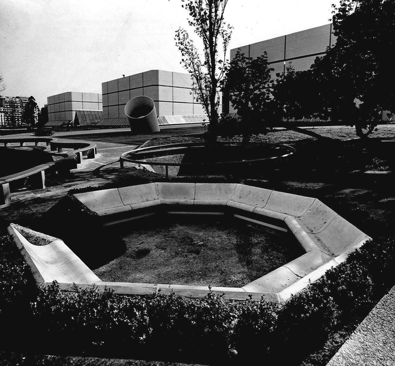 Octagonal concrete seating area with planted ground cover in a landscaped plaza with gridded facade beyond