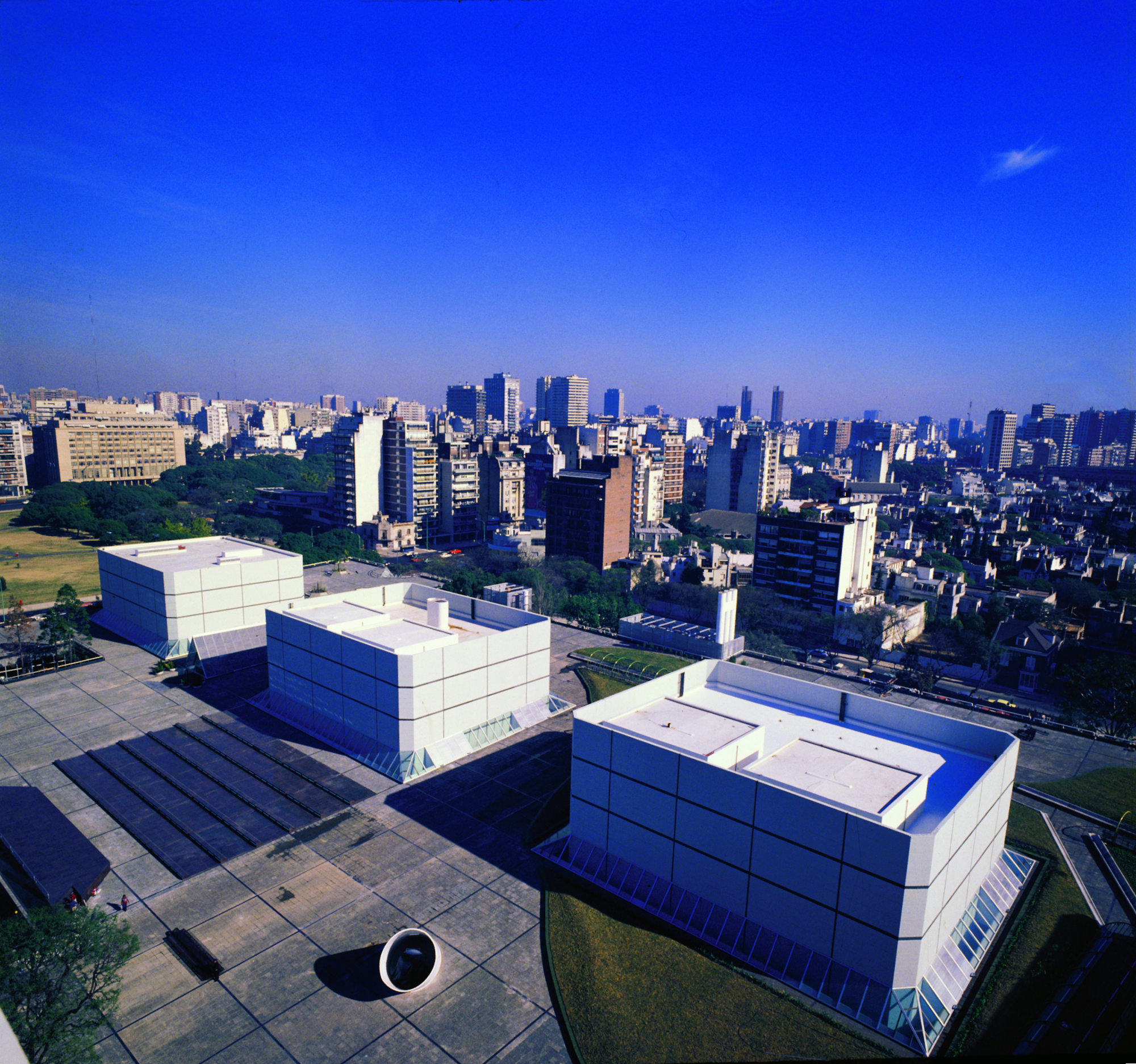 Aerial view of white paneled volumes with cylindrical sculpture on paved plaza overlooking dense urban skyline