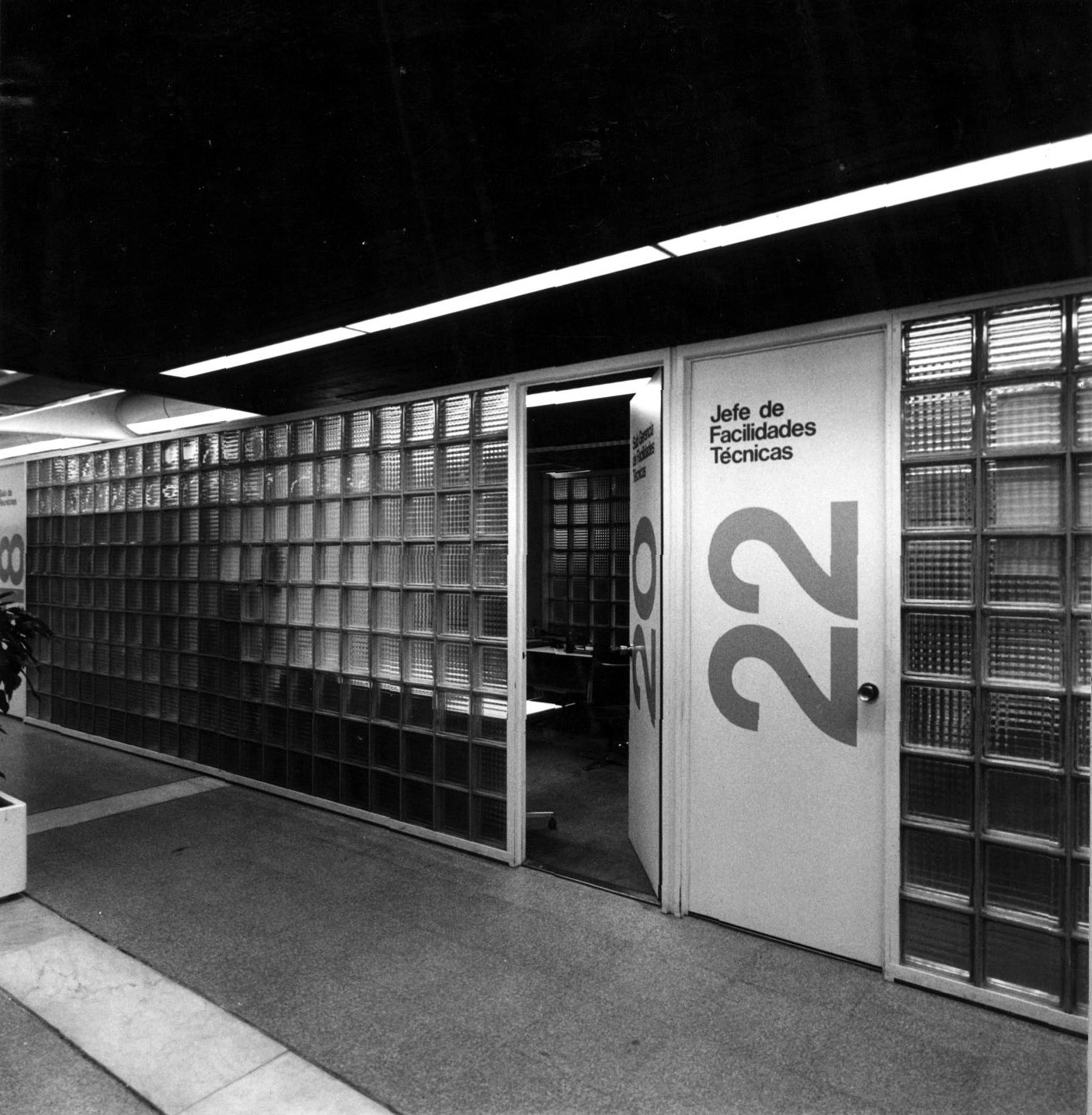 Corridor with glass block wall and door marked with numerals under linear ceiling light