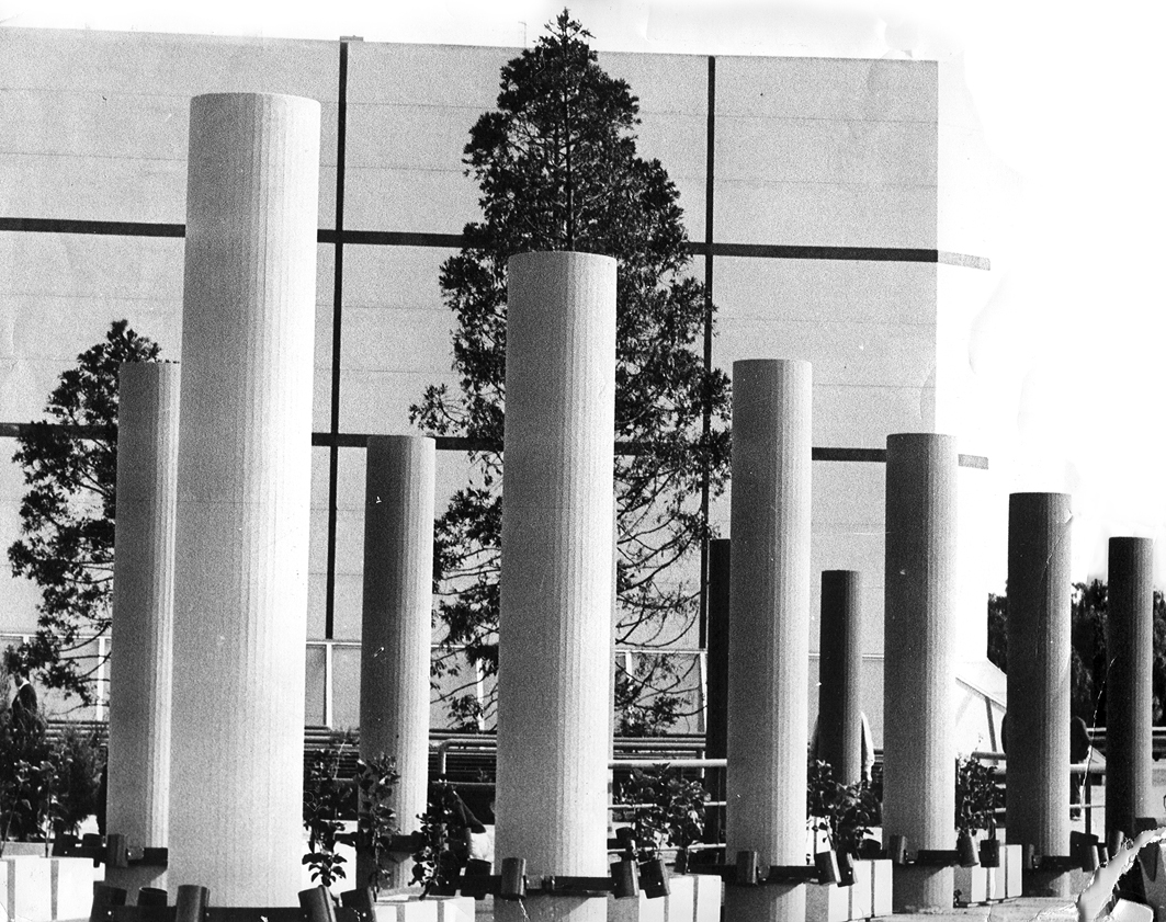 Grid of white cylindrical columns with horizontal bands and trees visible beyond