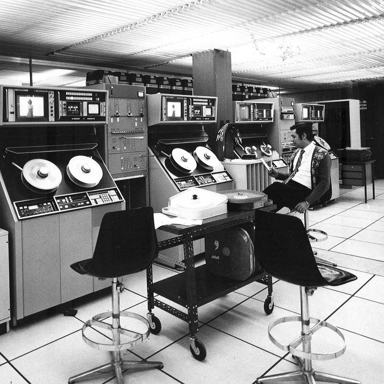 Computer room with reel-to-reel tape drives and operators seated at consoles beneath a ribbed ceiling