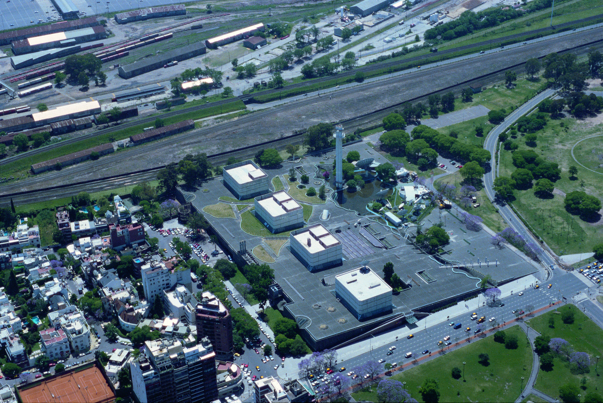 Aerial view of low-rise industrial buildings with white rooftop penthouses adjacent to railway tracks and parkland