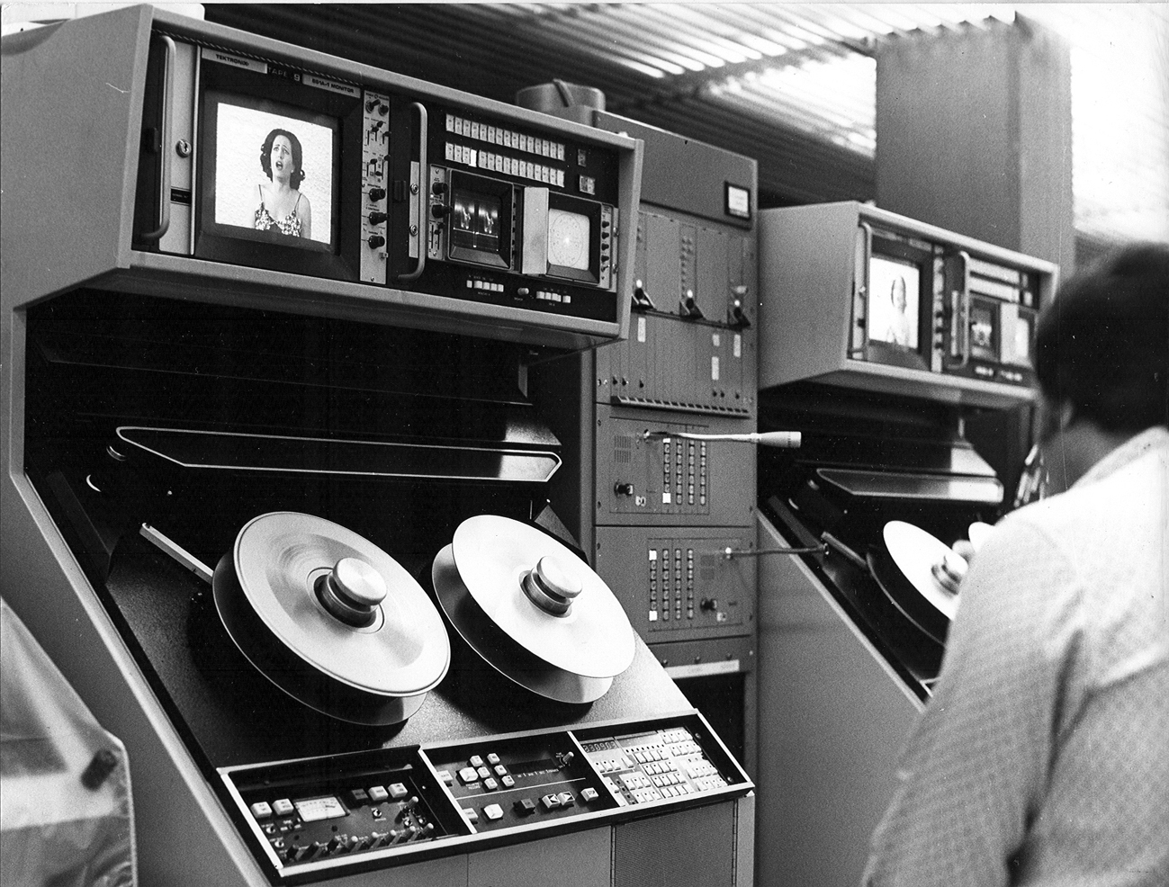 Operator working at a console with reel-to-reel tape drives and monitor screens in a ribbed ceiling room