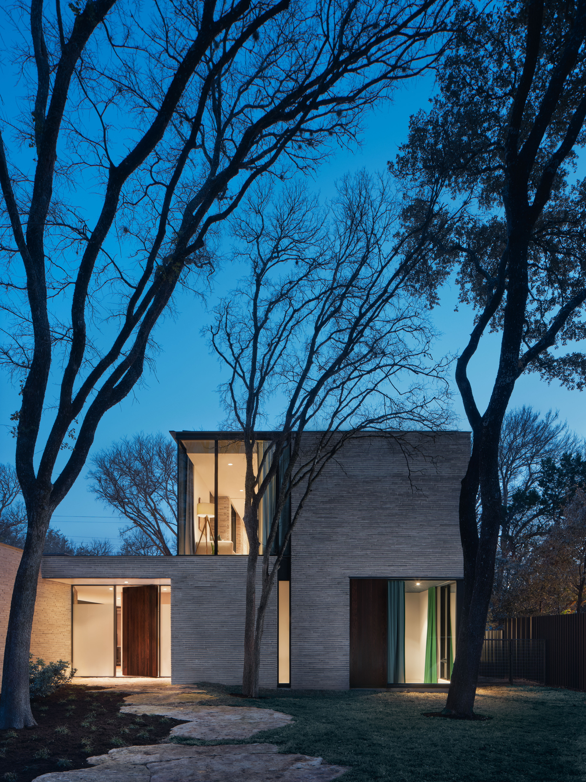 Brick facade with illuminated vertical window tower framed by bare winter trees at twilight