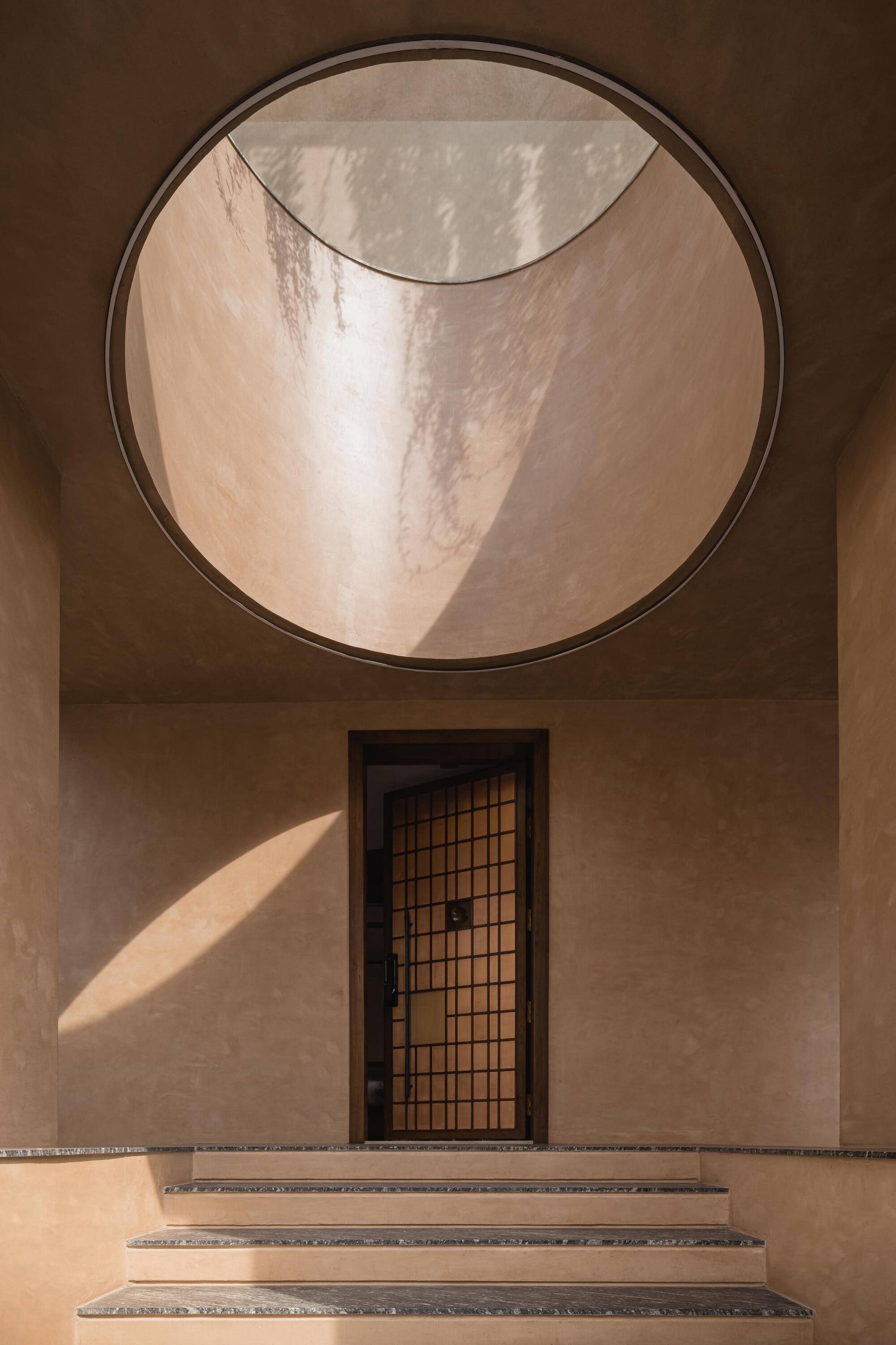 Entry steps leading to gridded door beneath circular skylight with curved plaster walls in afternoon light