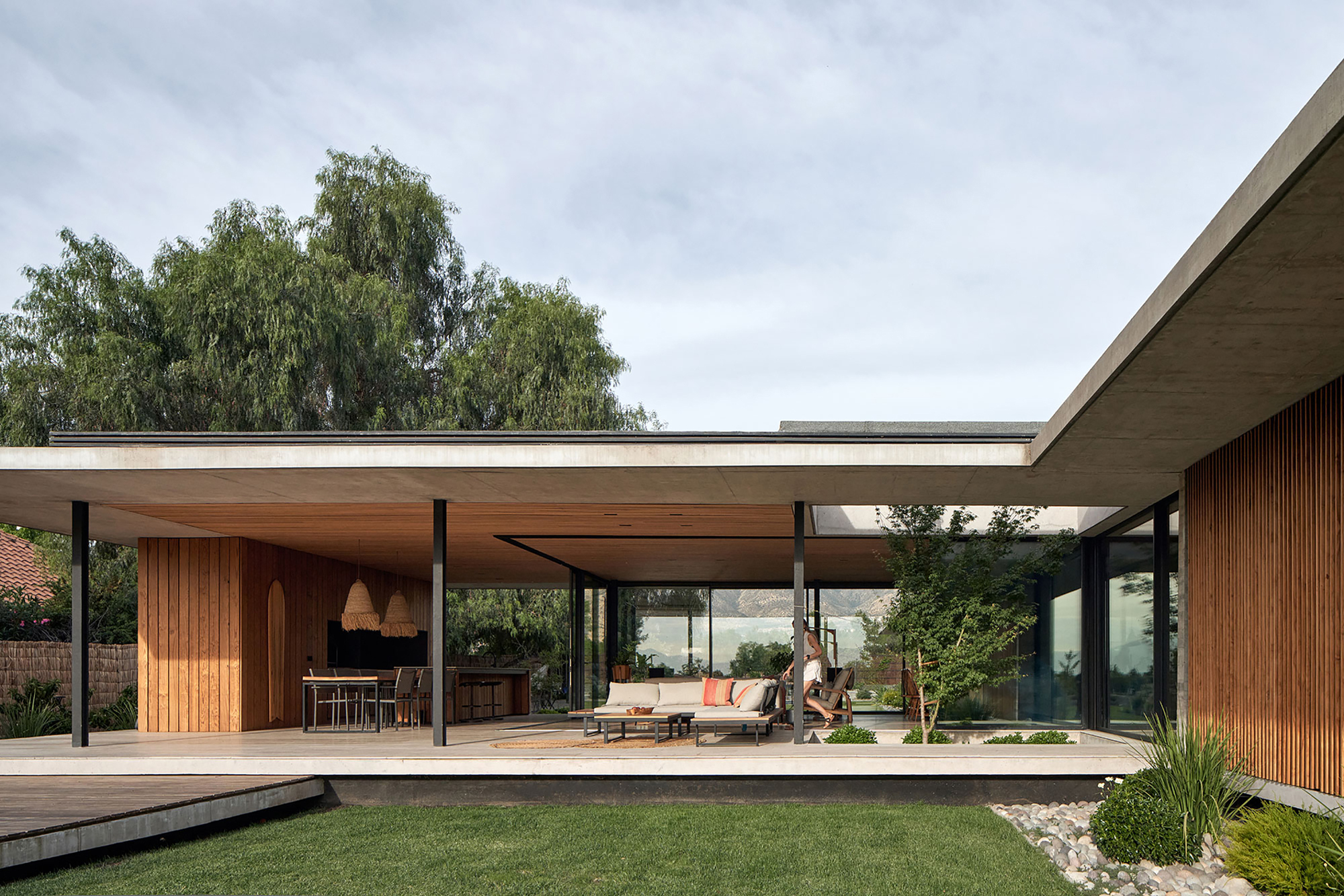 Covered terrace with timber ceiling and slender black columns sheltering outdoor seating and lawn