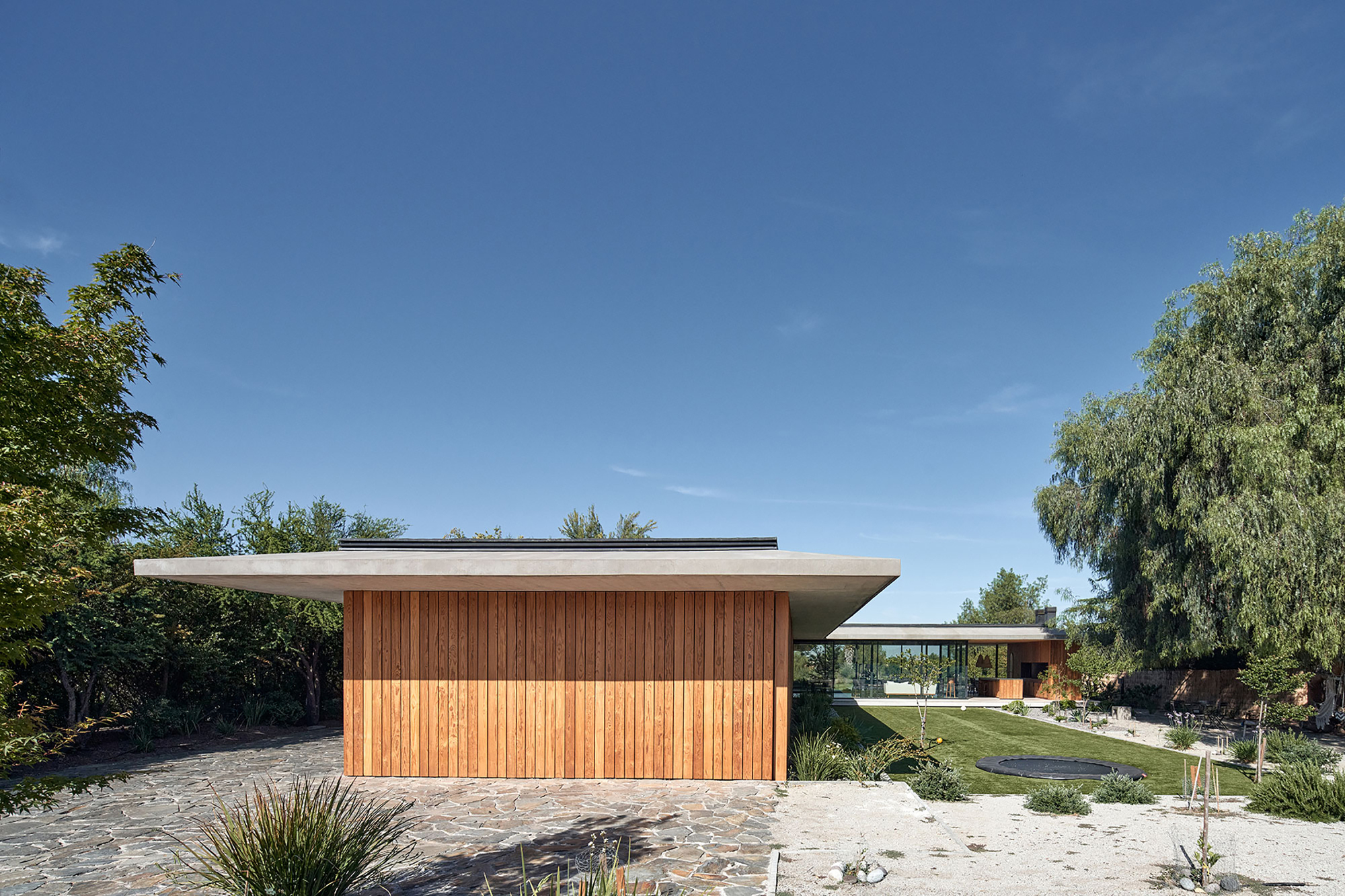 Street view of the timber-clad volume with extended roof plane and stone paving under clear skies
