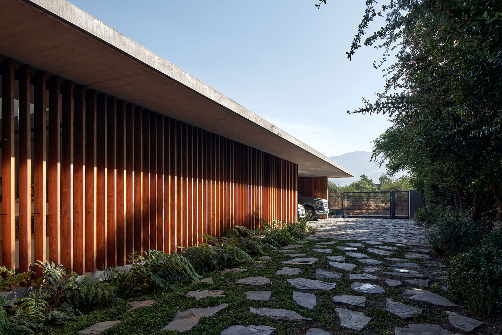Entry sequence with vertical timber battens casting shadows along stone paver path and carport beyond