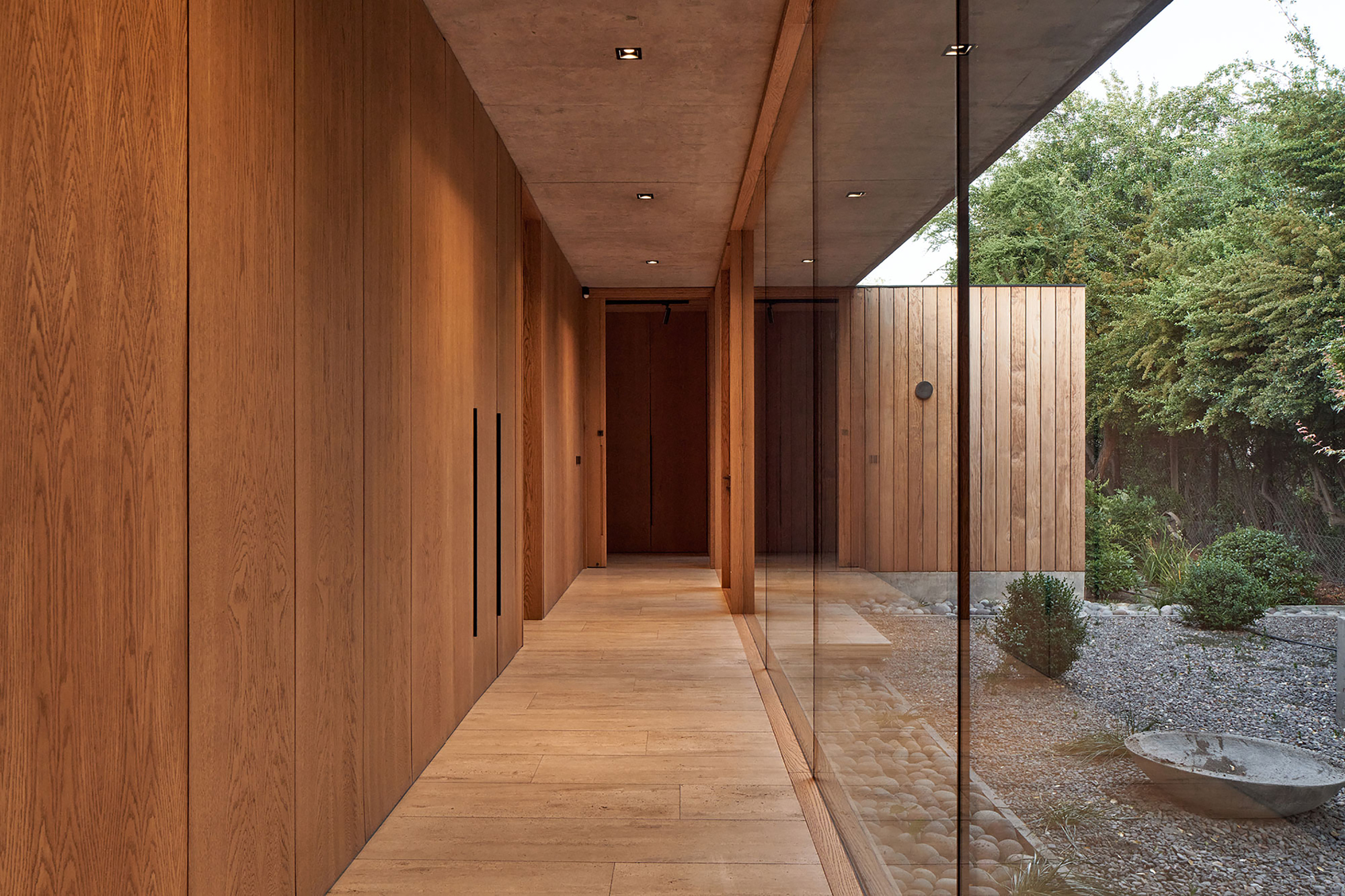 Timber-lined corridor with full-height glass wall overlooking a gravel courtyard with planted bowl and shrubs