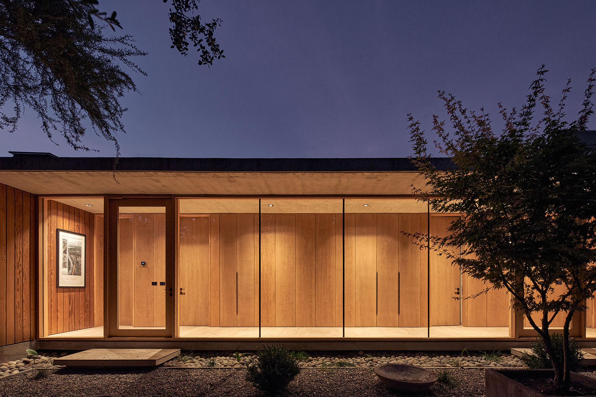 Illuminated timber facade with flat roof and glazed entry corridor at twilight beside planted gravel beds