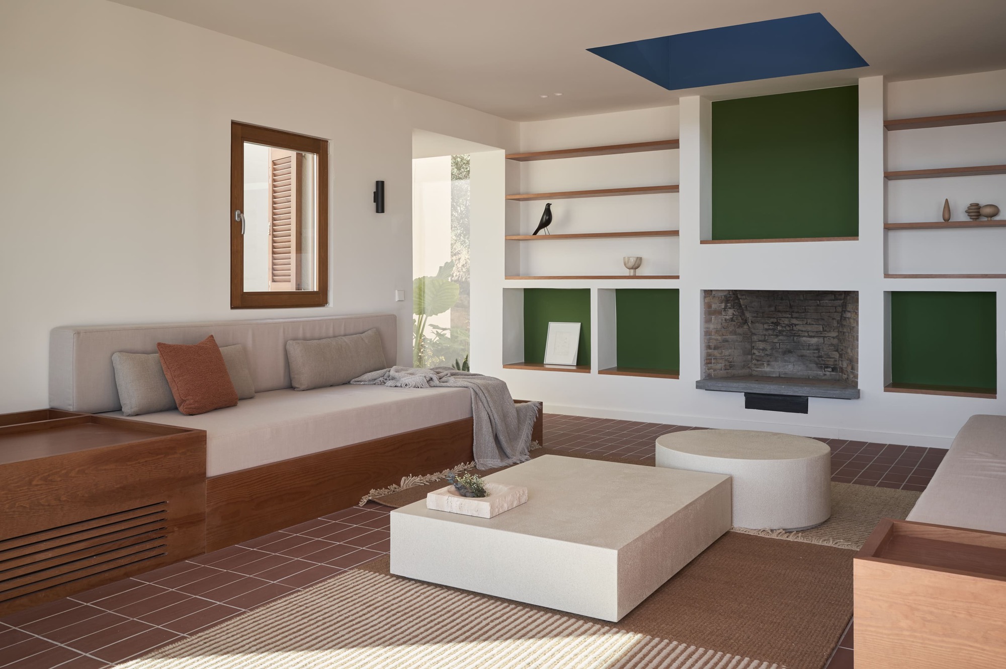 Living room with terracotta tile floor, built-in fireplace, green-painted niches and timber shelving under soft daylight