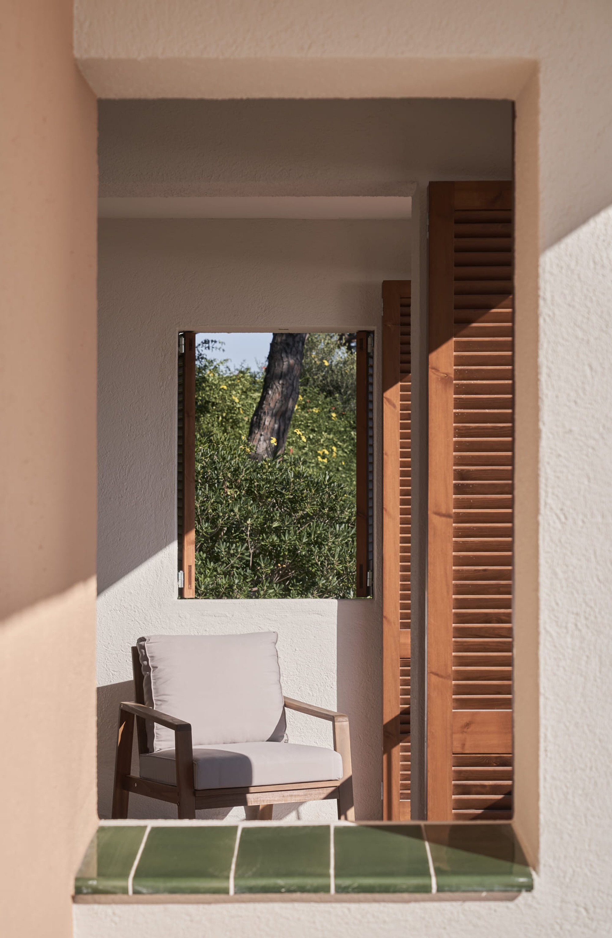 Small balcony with timber louvered shutters, green tile threshold, chair and framed view of tree canopy