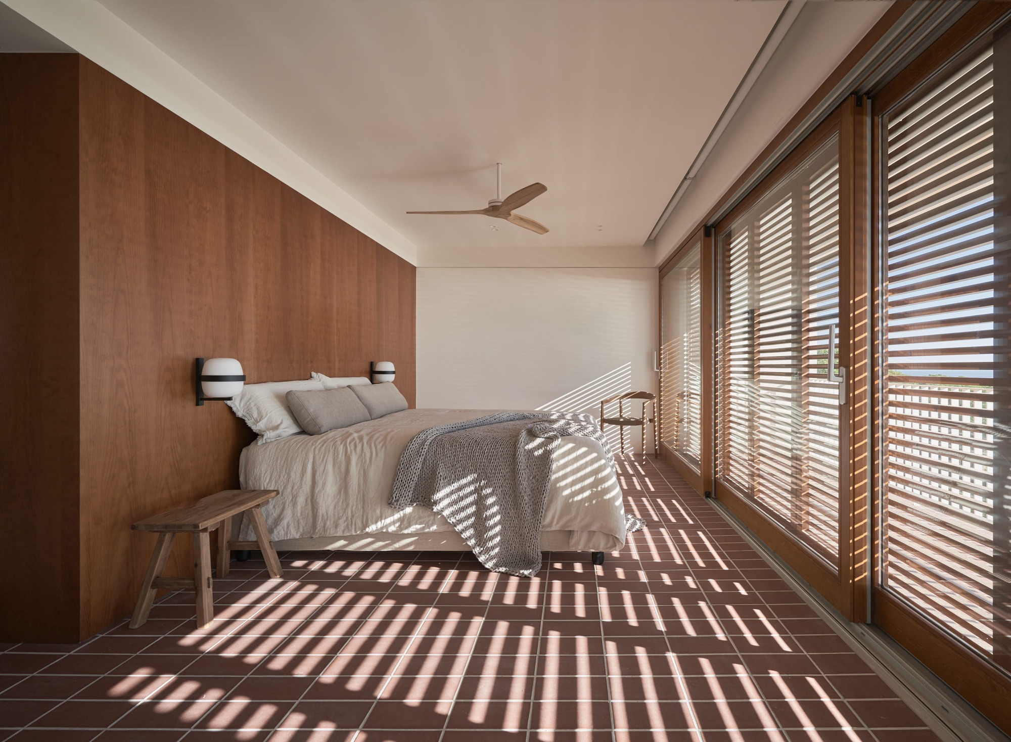 Bedroom with timber wall paneling and louvered shutters casting striped shadows across terracotta tile floor