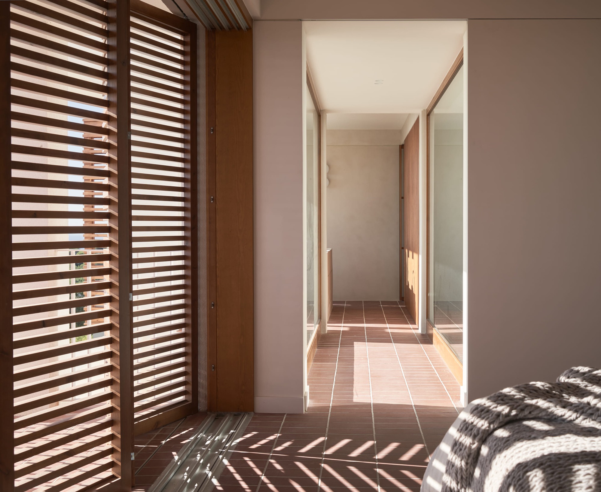 Corridor with timber louvered doors filtering sunlight into geometric shadow patterns on terracotta tile and pink walls
