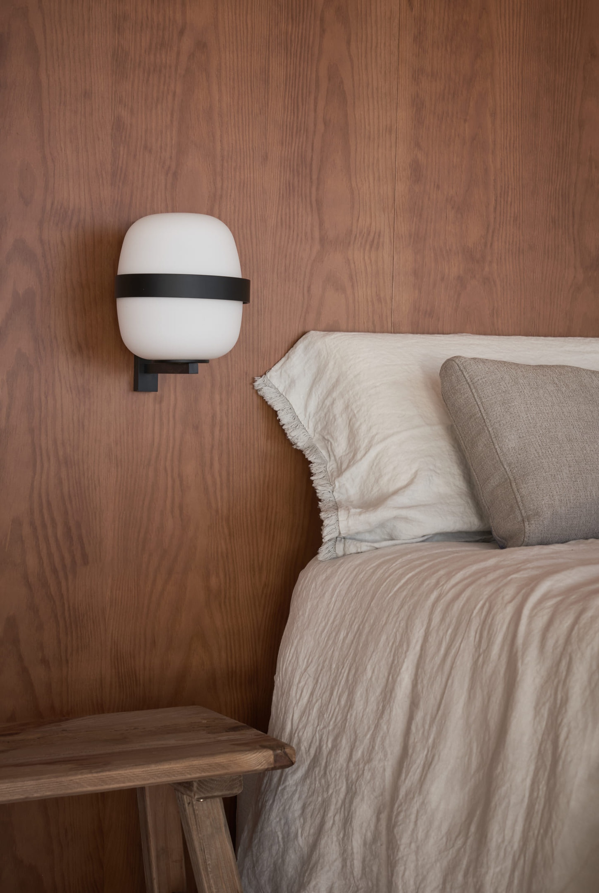 Bedroom wall clad in vertical wood grain panels with a wall-mounted white sconce above linen bedding