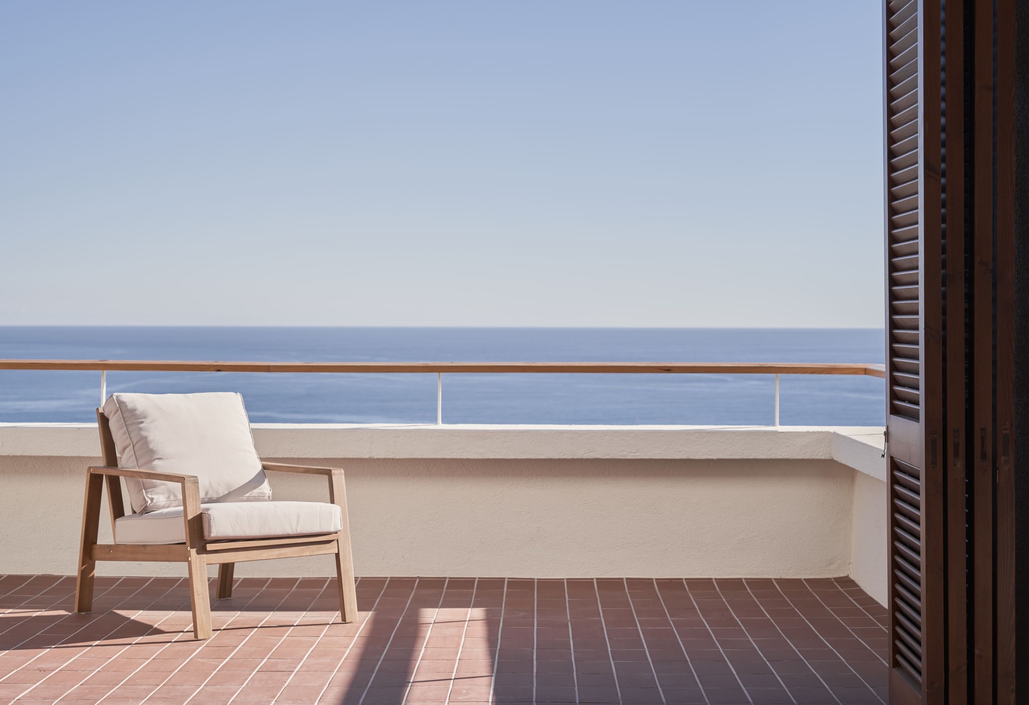 Terracotta-tiled terrace with timber chair overlooking the ocean through a glass and metal railing