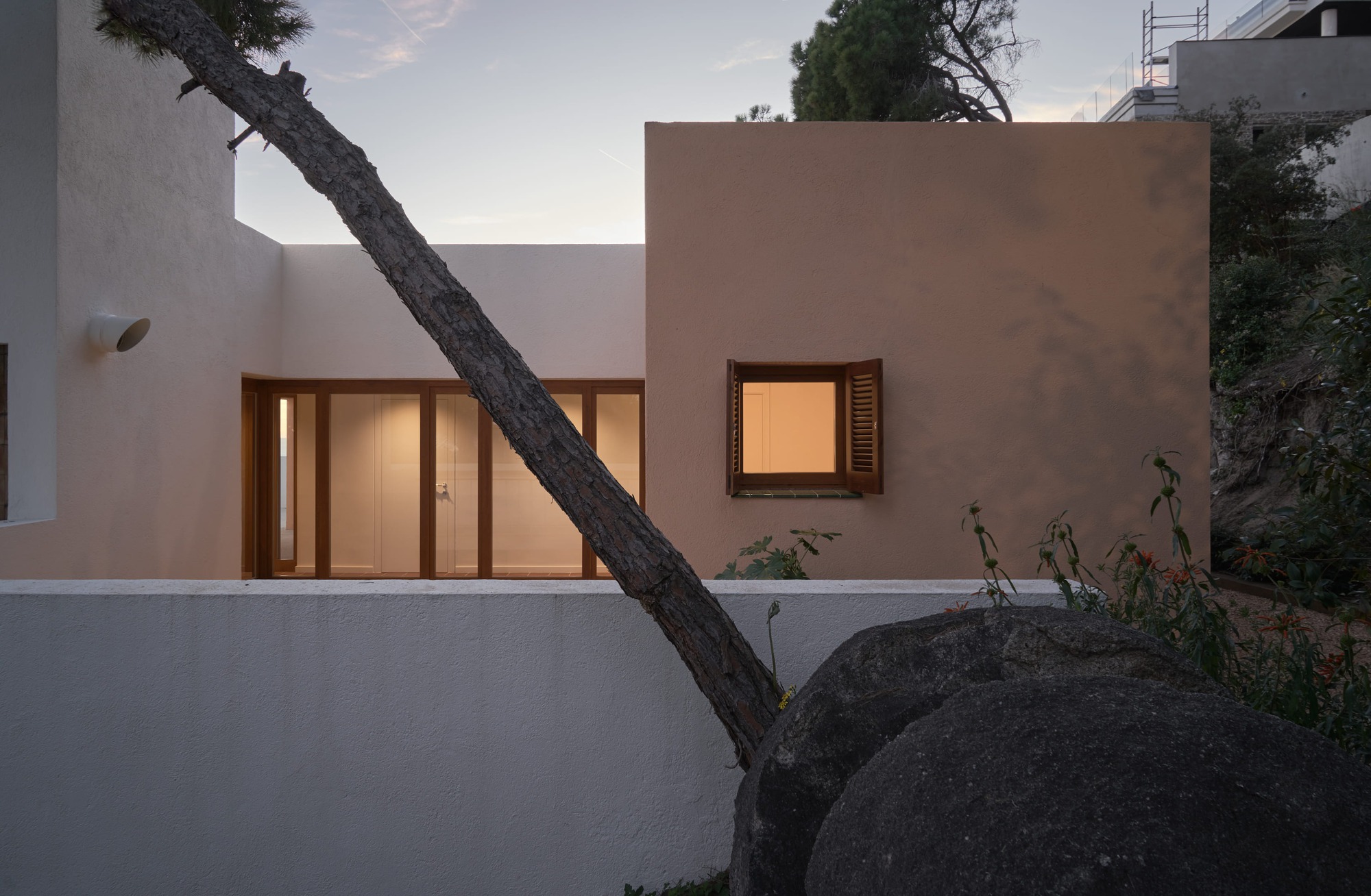 White stucco facade with illuminated window opening at dusk and a leaning tree trunk