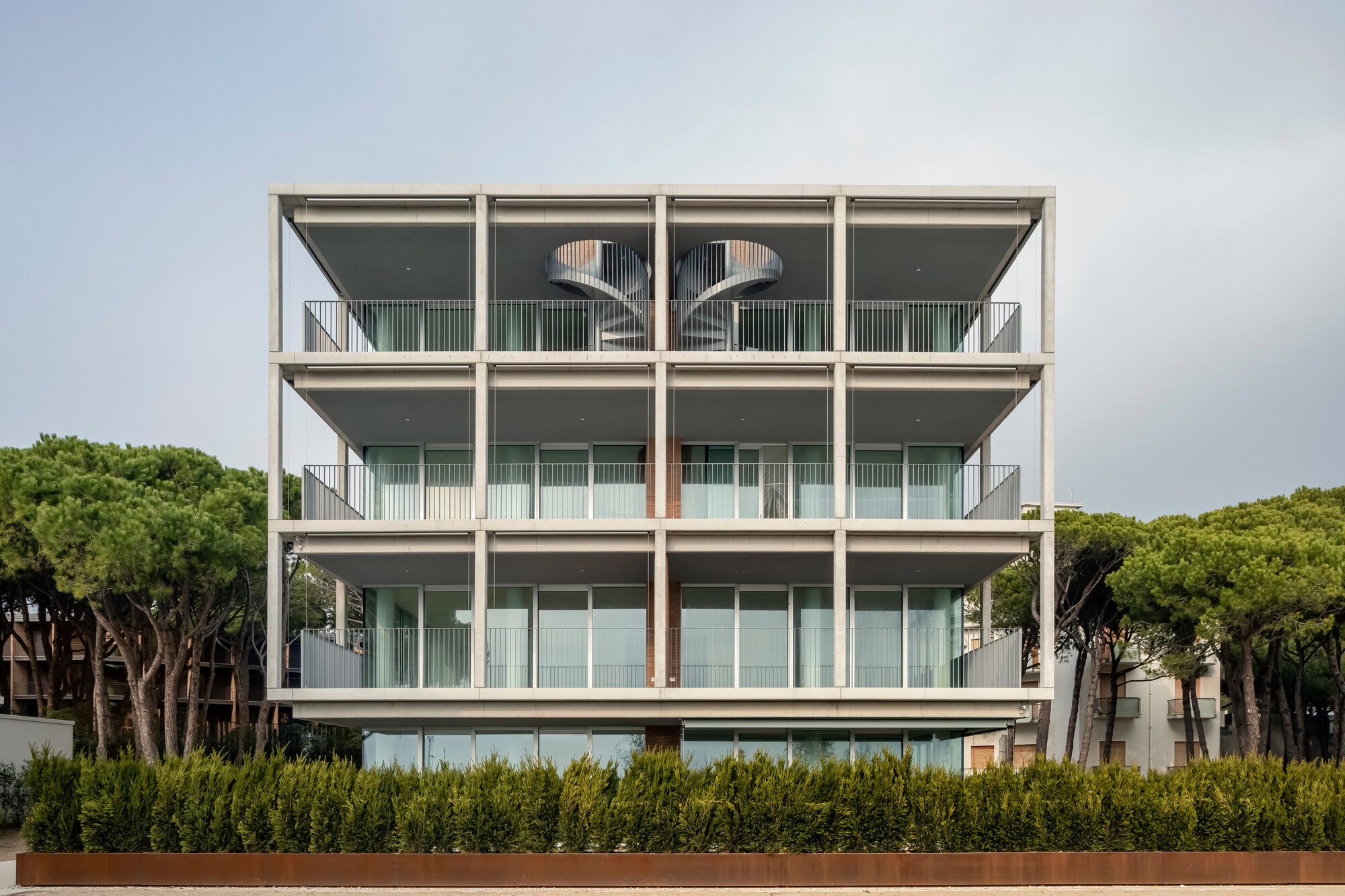 Frontal view of the four-storey concrete frame facade with open balconies behind a hedge of green shrubs
