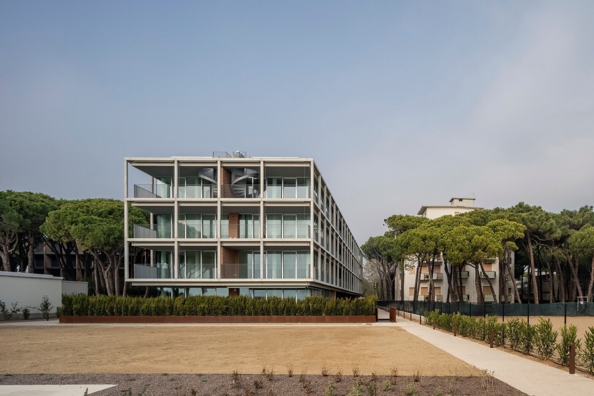 Oblique view across the sand courtyard showing the building flanked by dense pine groves under grey sky