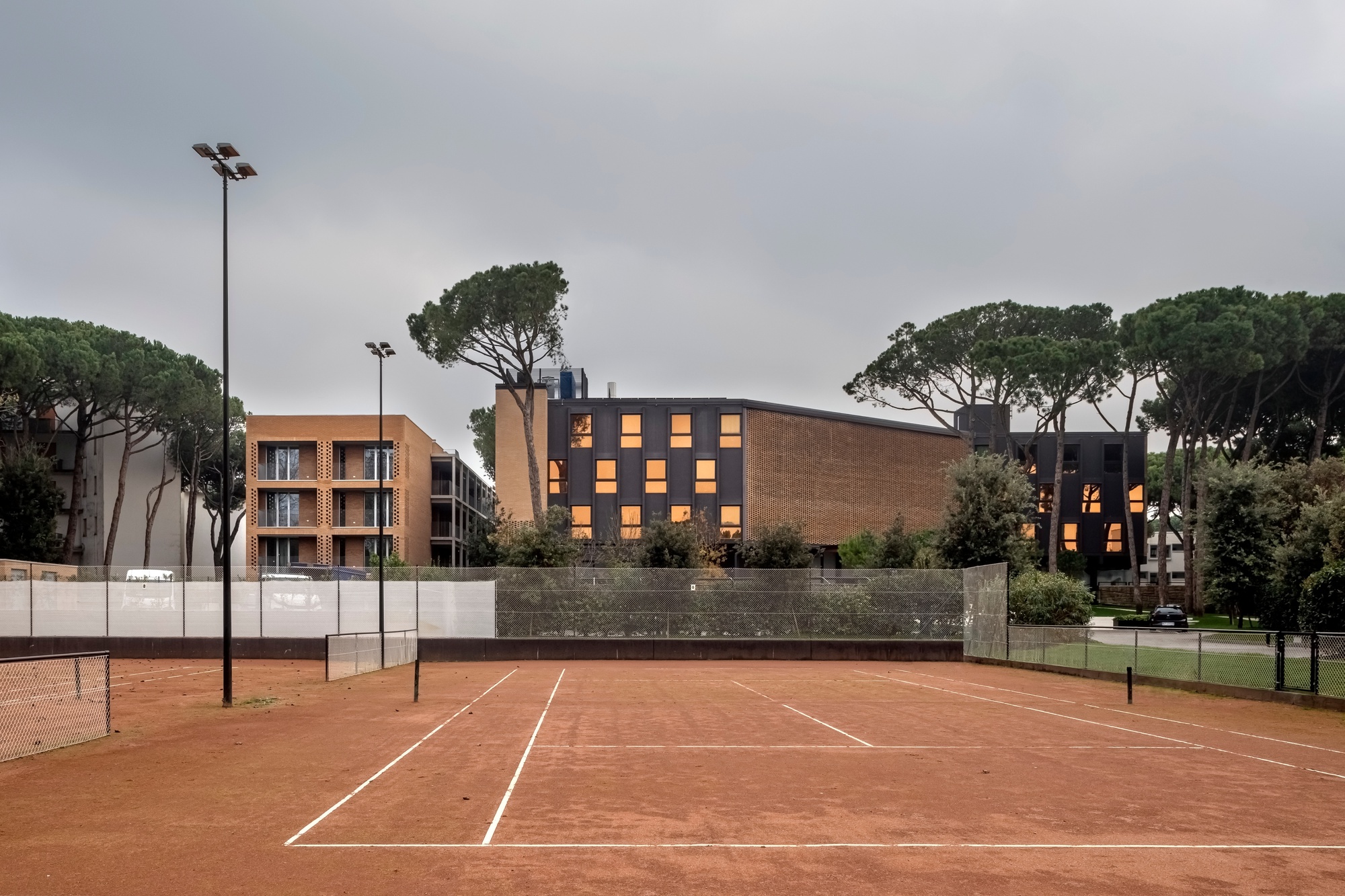 Perforated brick facade with illuminated windows viewed across a red clay tennis court at dusk