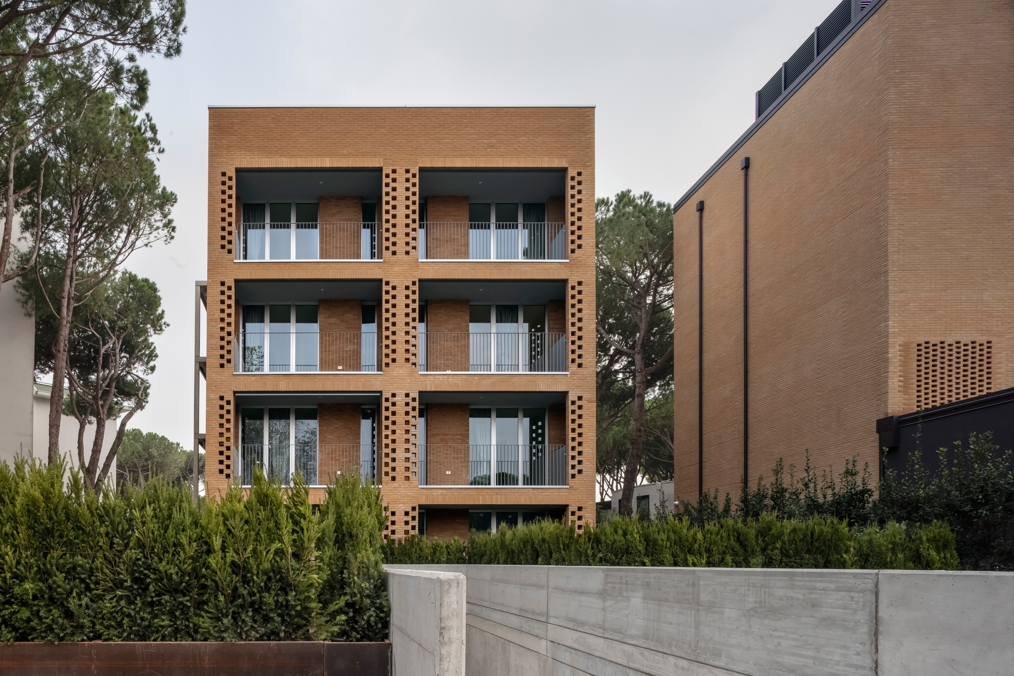 Four-story perforated brick facade with recessed balconies behind a concrete retaining wall and planted hedge