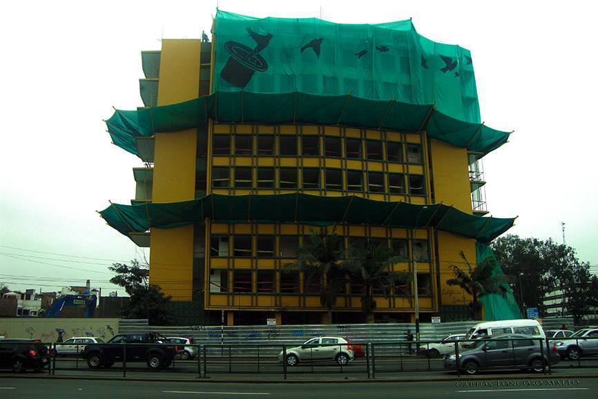 Yellow facade wrapped in green construction netting with parked vehicles visible along the street below
