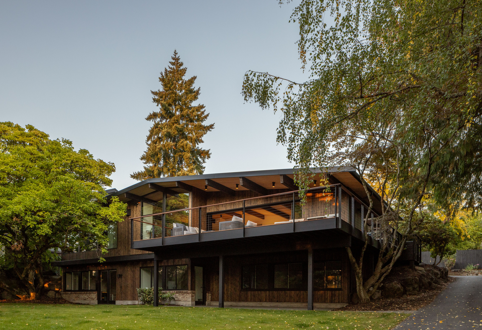 Two-storey residence with timber cladding and exposed roof beams surrounded by mature trees at dusk