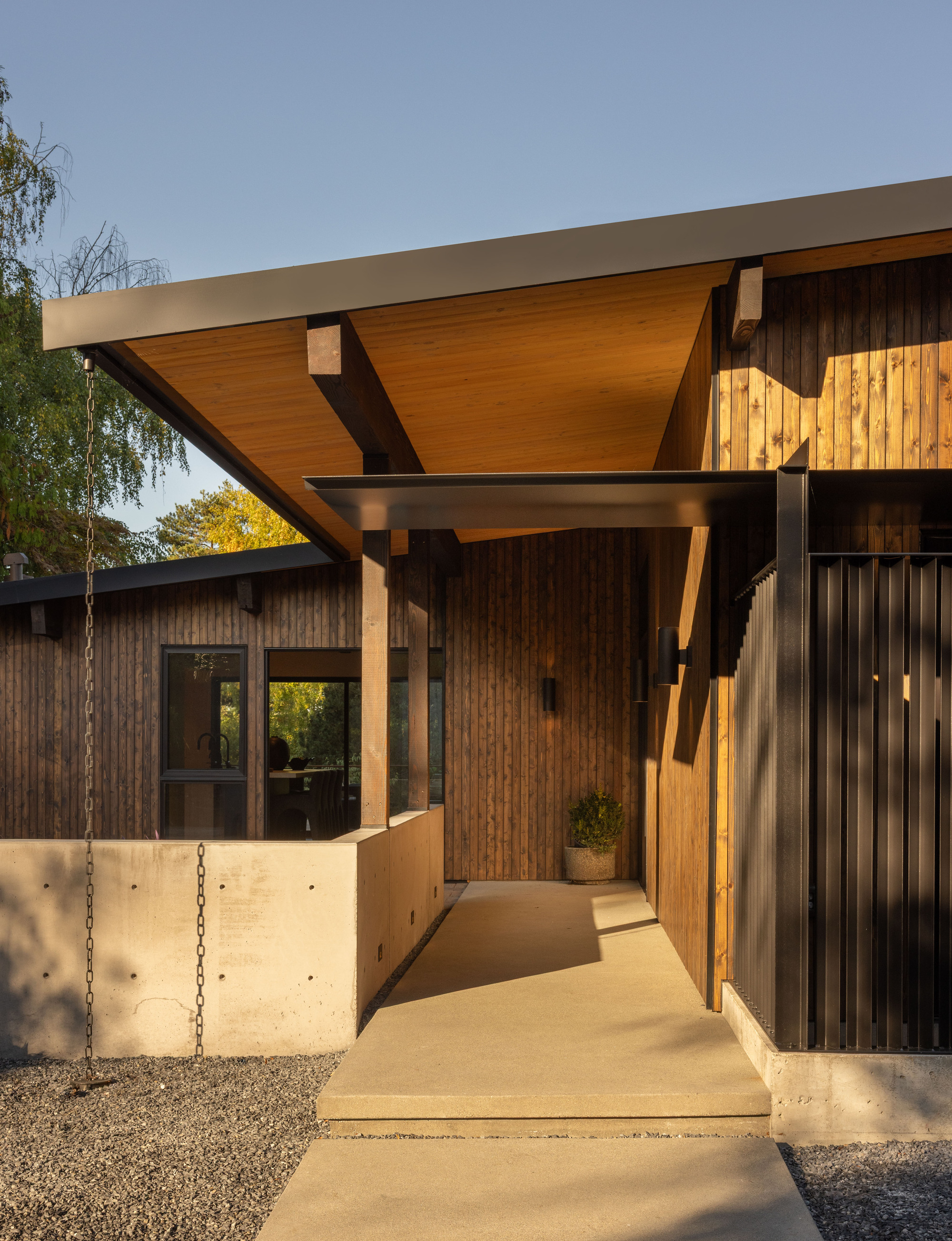 Entry canopy with exposed timber soffit and diagonal bracing above a concrete walkway at sunset