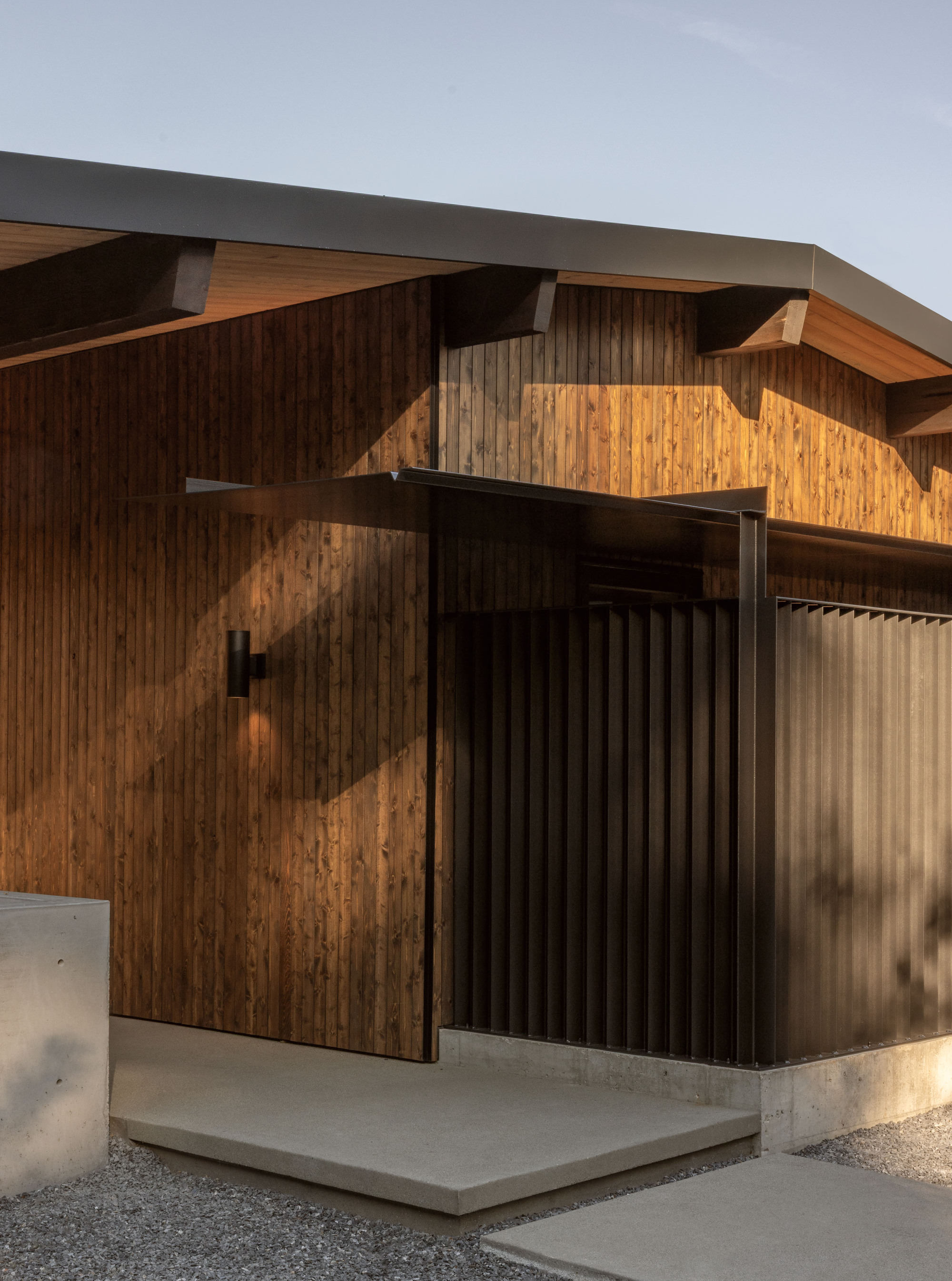 Entry elevation showing vertical timber siding with diagonal sunlight patterns and concrete plinth below