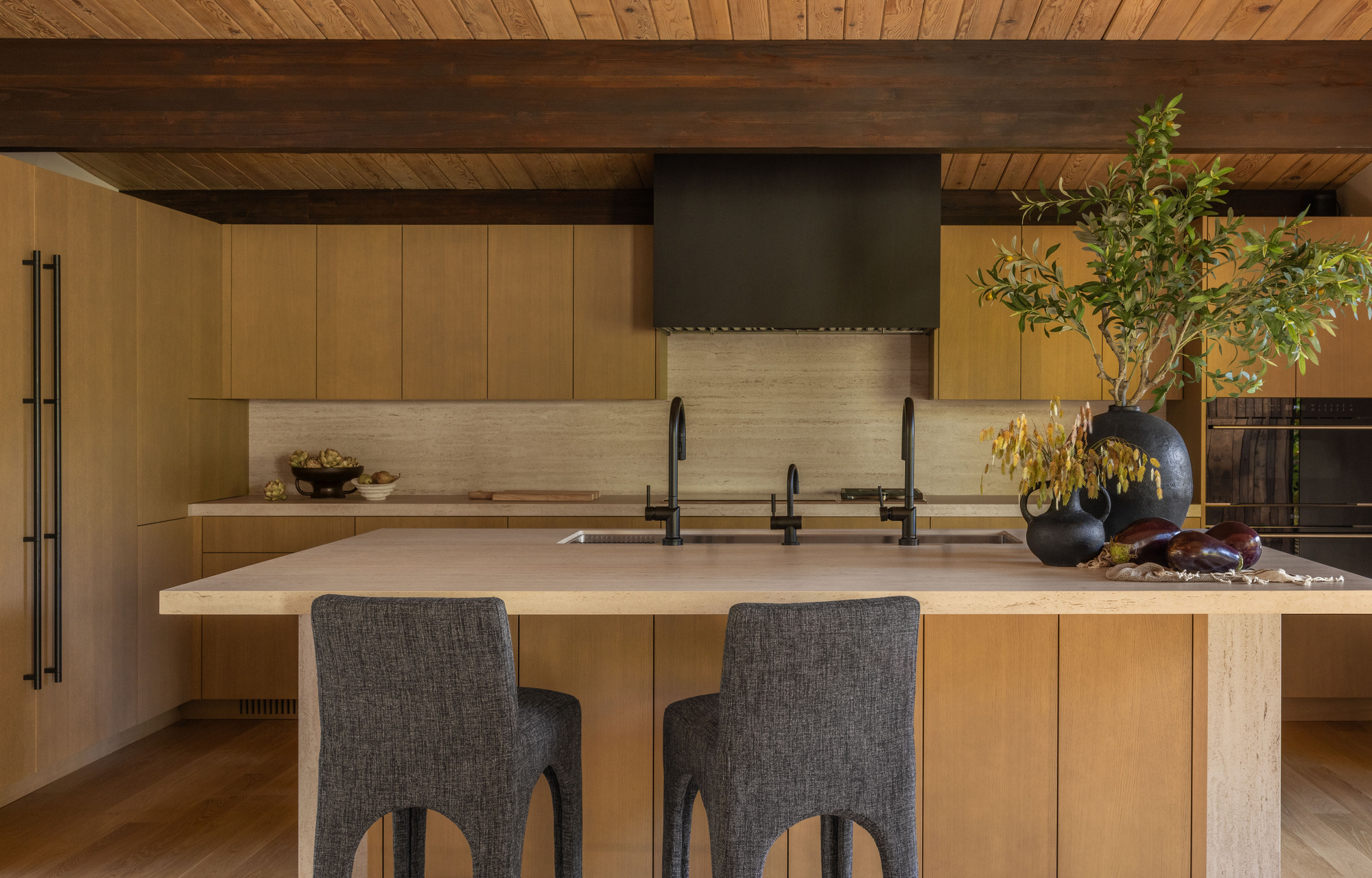 Kitchen island with stone countertop and dark grey upholstered stools beneath exposed timber beams