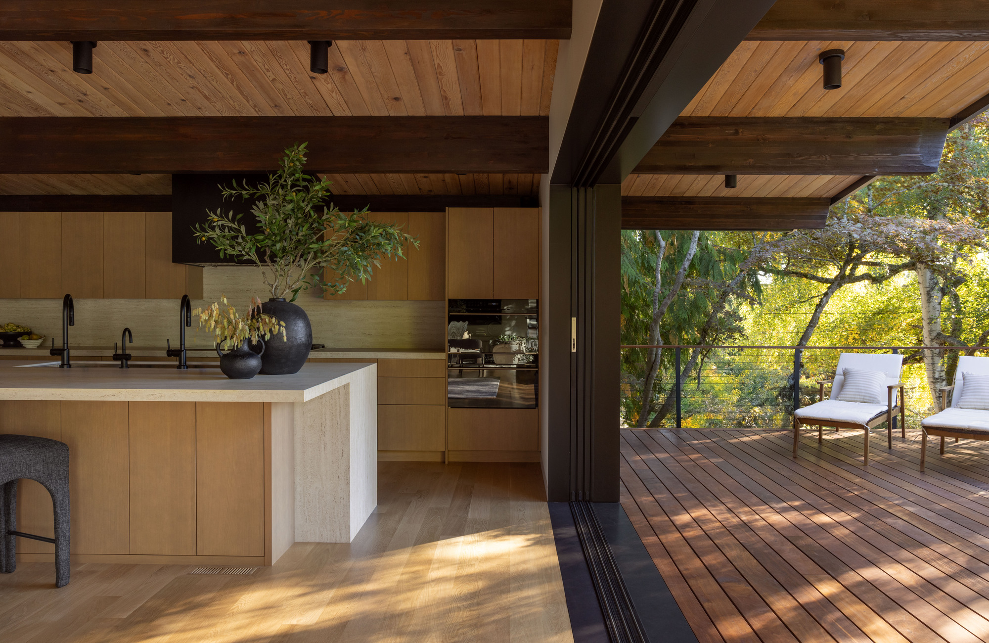 Open kitchen threshold leading to timber deck with lounge chairs framed by tree canopy