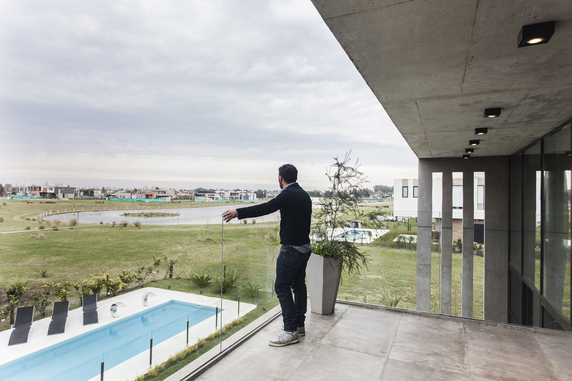 Man standing on a concrete terrace with glass balustrade, gesturing toward a pool and open lawn under an overcast sky
