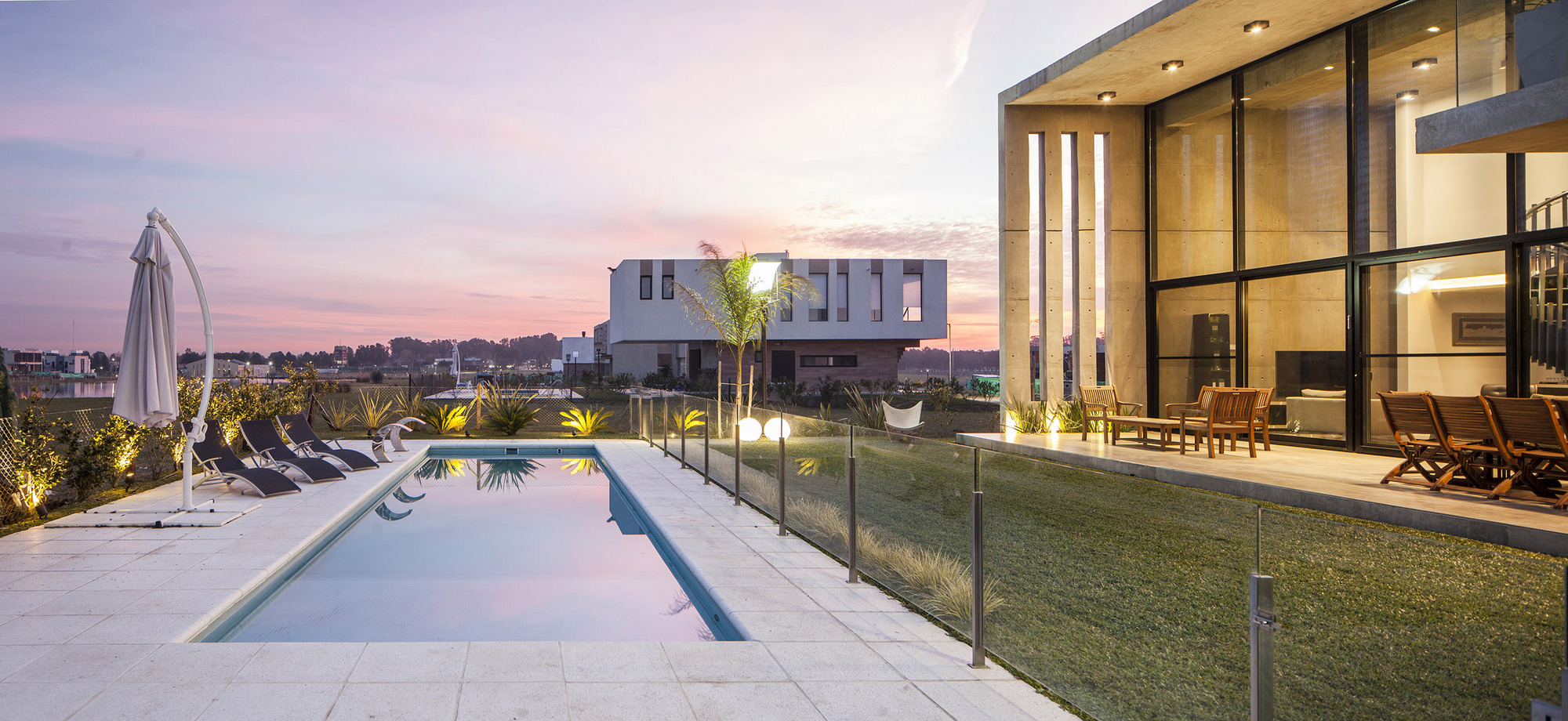 Rectangular swimming pool with lounge chairs and a folded umbrella beside a glazed concrete house at pink dusk
