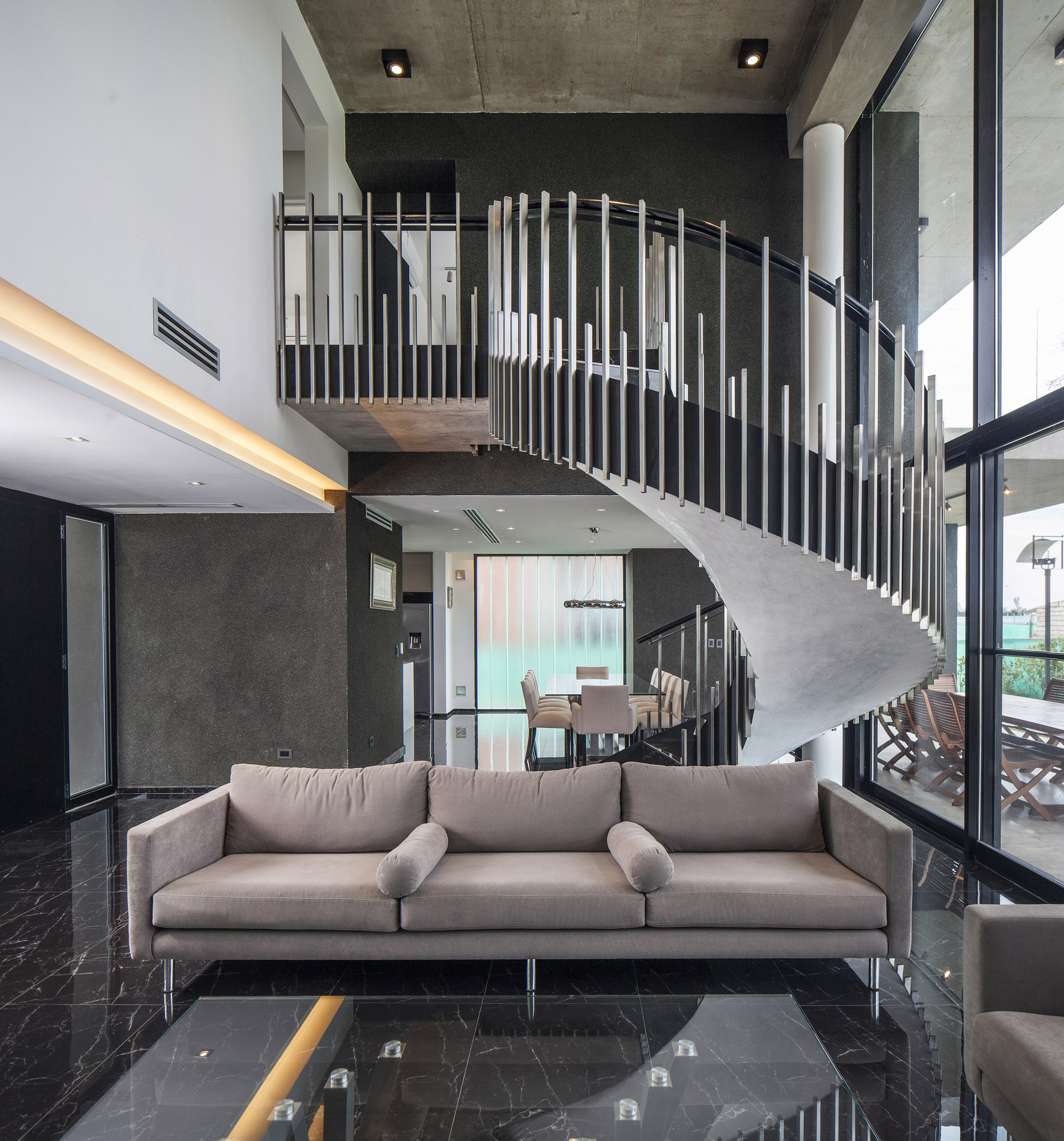 Spiral concrete staircase with slender vertical steel balusters rising behind a grey sofa on a black marble floor